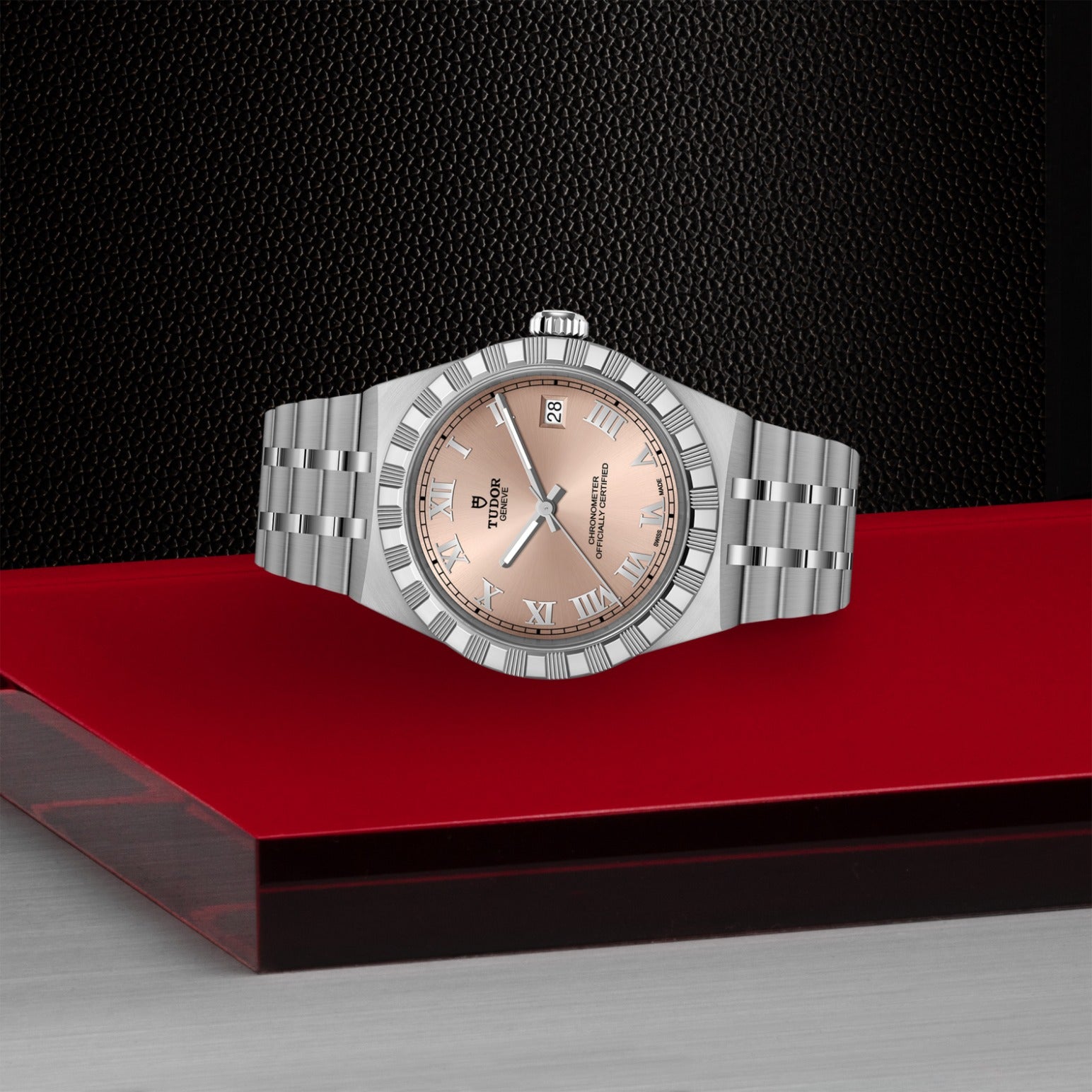 The TUDOR Royal wristwatch by TUDOR features a silver case, fluted bezel, and salmon dial with Roman numerals and date window, displayed on a red and gray geometric surface against a textured black background.