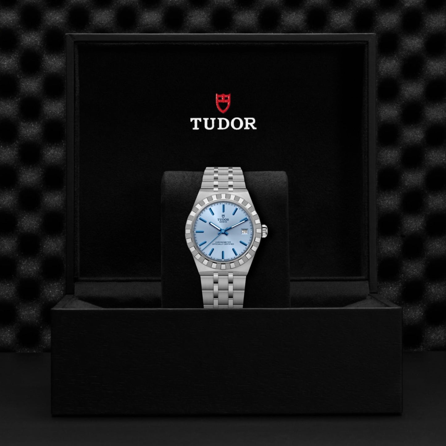 A TUDOR Royal wristwatch in stainless steel with a light blue dial rests on a black cushion inside an open black TUDOR box, set against a dark textured background—a refined example of sport-chic design.