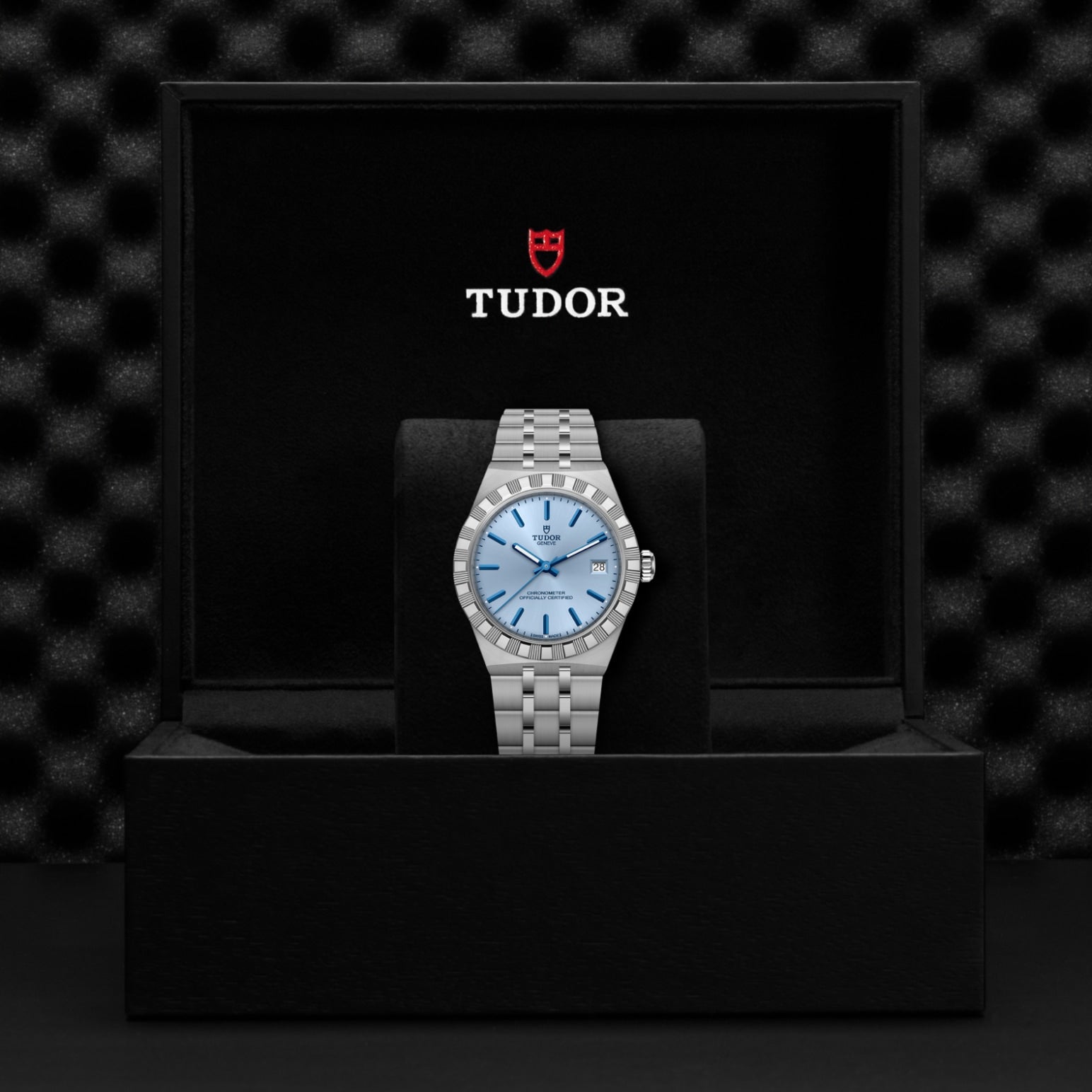 A TUDOR Royal wristwatch in stainless steel with a light blue dial rests on a black cushion inside an open black TUDOR box, set against a dark textured background—a refined example of sport-chic design.