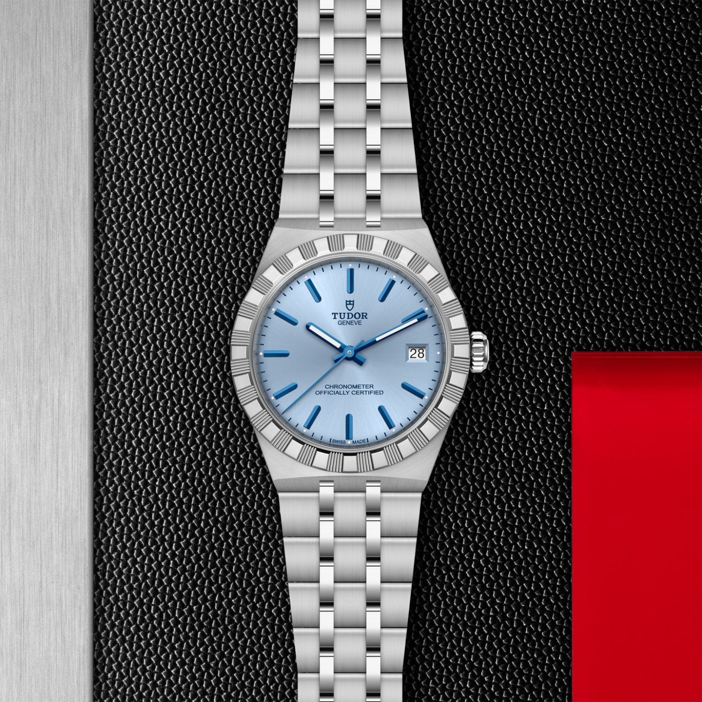 A TUDOR Royal wristwatch by TUDOR, featuring a silver finish, light blue dial with date at 3 o’clock, and stainless steel bracelet, rests on a black and metallic surface with red accents—showcasing refined sport-chic style.