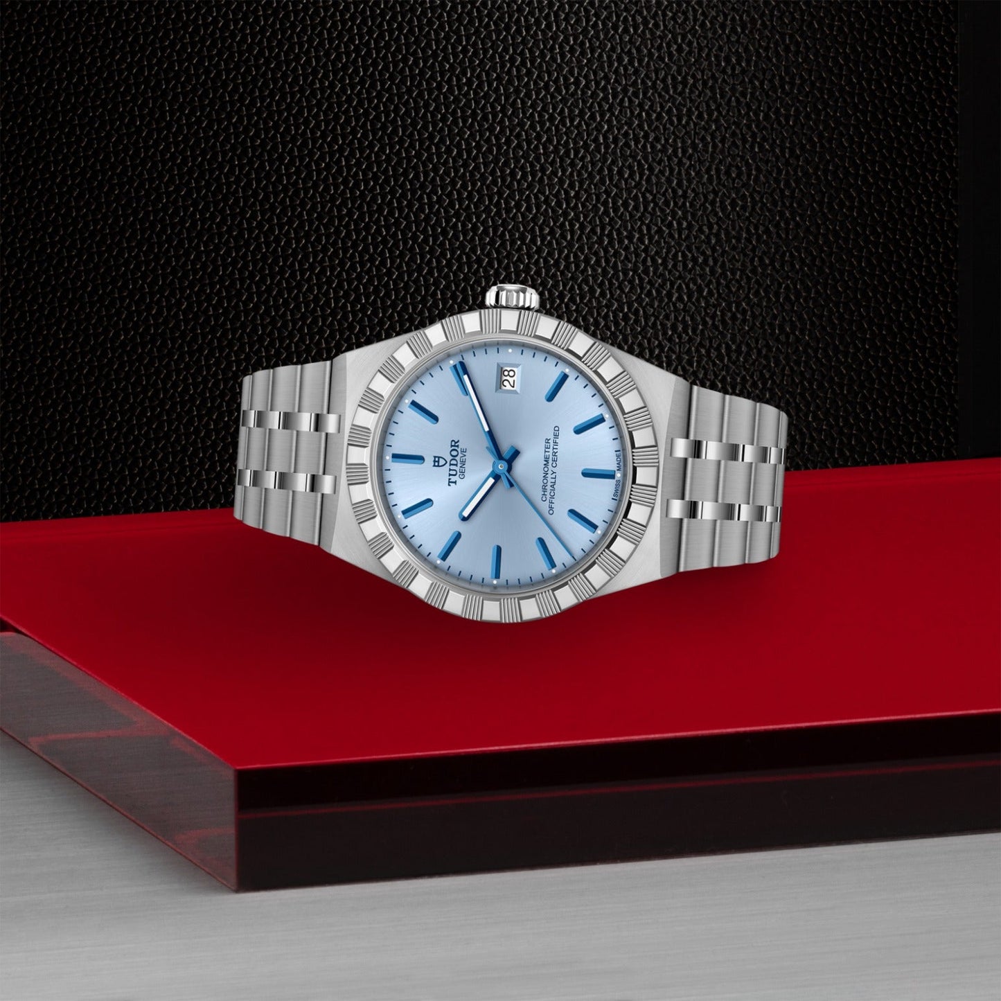 A TUDOR Royal wristwatch by TUDOR, featuring a silver stainless steel case and light blue dial with date display, is displayed on a red and gray geometric surface against a textured black background.