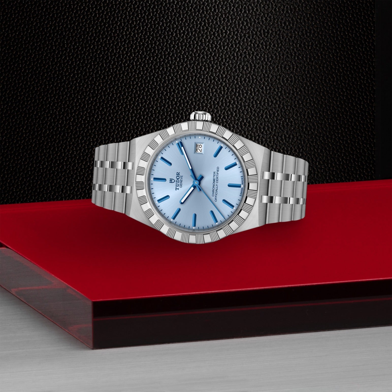 A TUDOR Royal wristwatch by TUDOR, featuring a silver stainless steel case and light blue dial with date display, is displayed on a red and gray geometric surface against a textured black background.