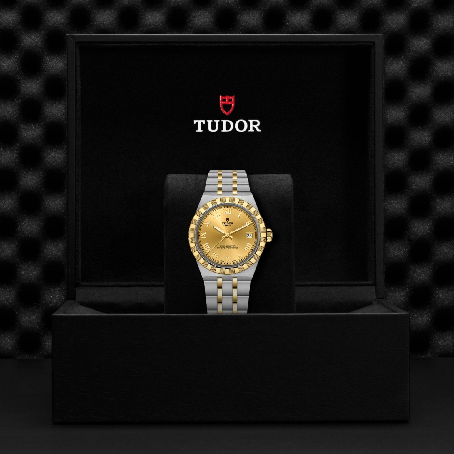A TUDOR Royal two-tone wristwatch with a gold face and silver & gold bracelet rests in an open black TUDOR-branded watch box with a soft, textured interior, reflecting the automatic sport-chic legacy of the TUDOR Royal.