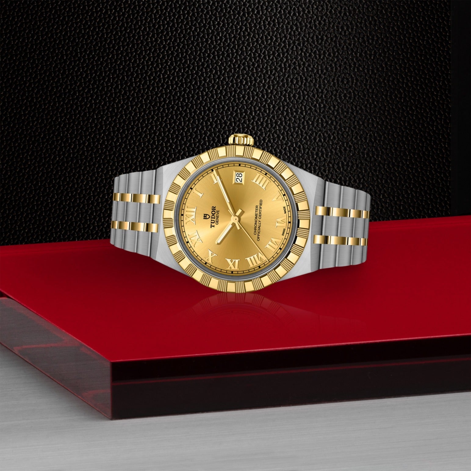 The TUDOR Royal by TUDOR, featuring a gold and silver design with a gold dial and Roman numerals, sits on a glossy red surface against a textured black backdrop—a refined automatic sport-chic wristwatch.