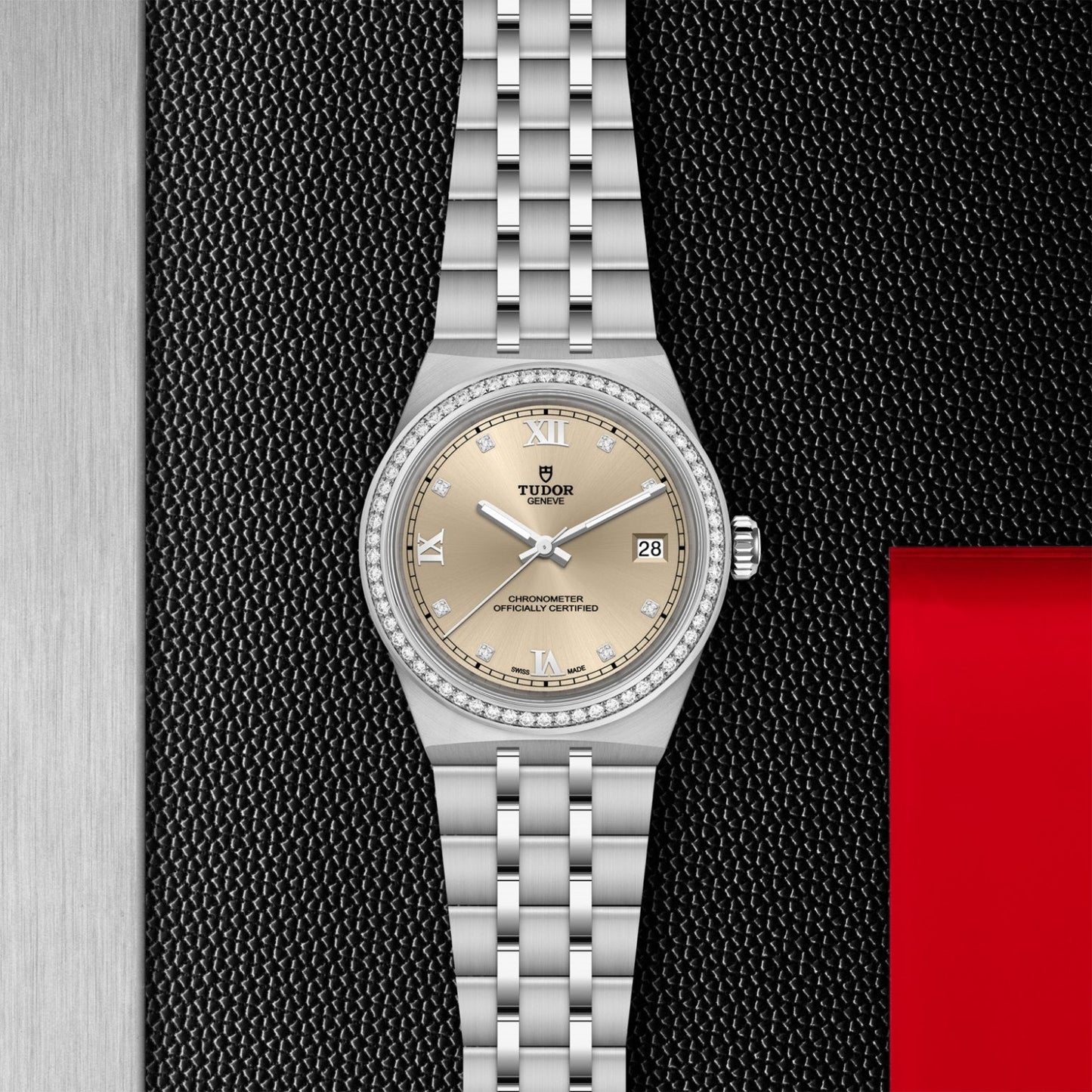 The TUDOR Royal wristwatch features a silver metal bracelet, diamond-set bezel, champagne dial with Roman numerals, and a date window at 3 o’clock, elegantly showcased on a black and metallic textured background.