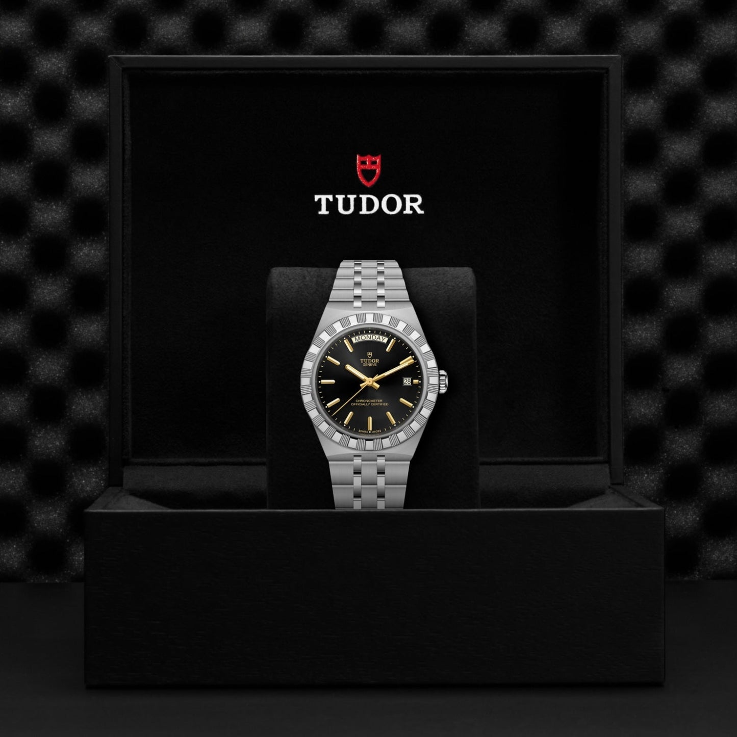 A TUDOR Royal stainless steel watch with a black dial and silver bracelet is presented in an open black box, featuring the TUDOR logo and name on the inside lid against a textured background.