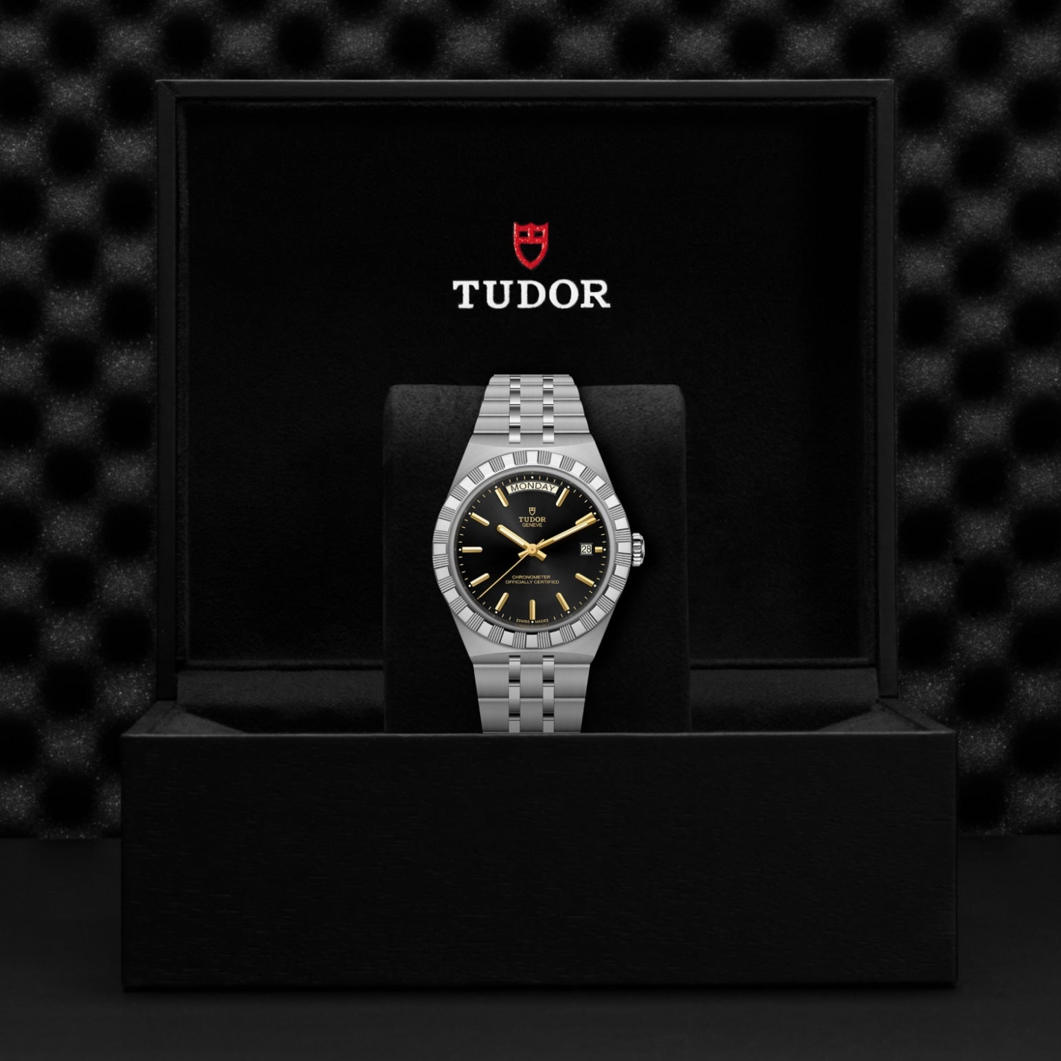 A TUDOR Royal stainless steel watch with a black dial and silver bracelet is presented in an open black box, featuring the TUDOR logo and name on the inside lid against a textured background.
