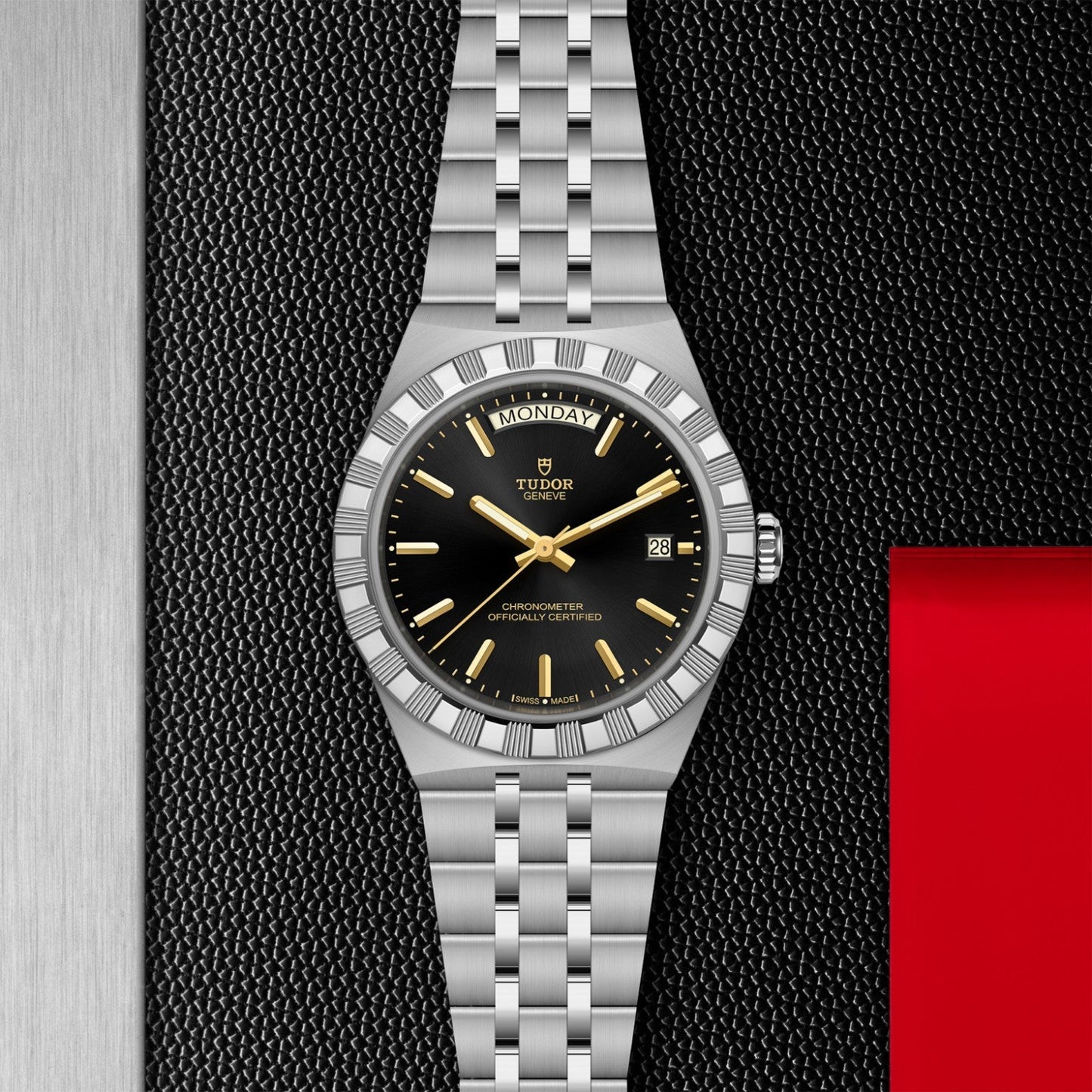 The TUDOR Royal by TUDOR, featuring a black dial, silver hour markers, day-date display, and stainless steel bracelet, epitomizes sport-chic style on a textured black and red background.