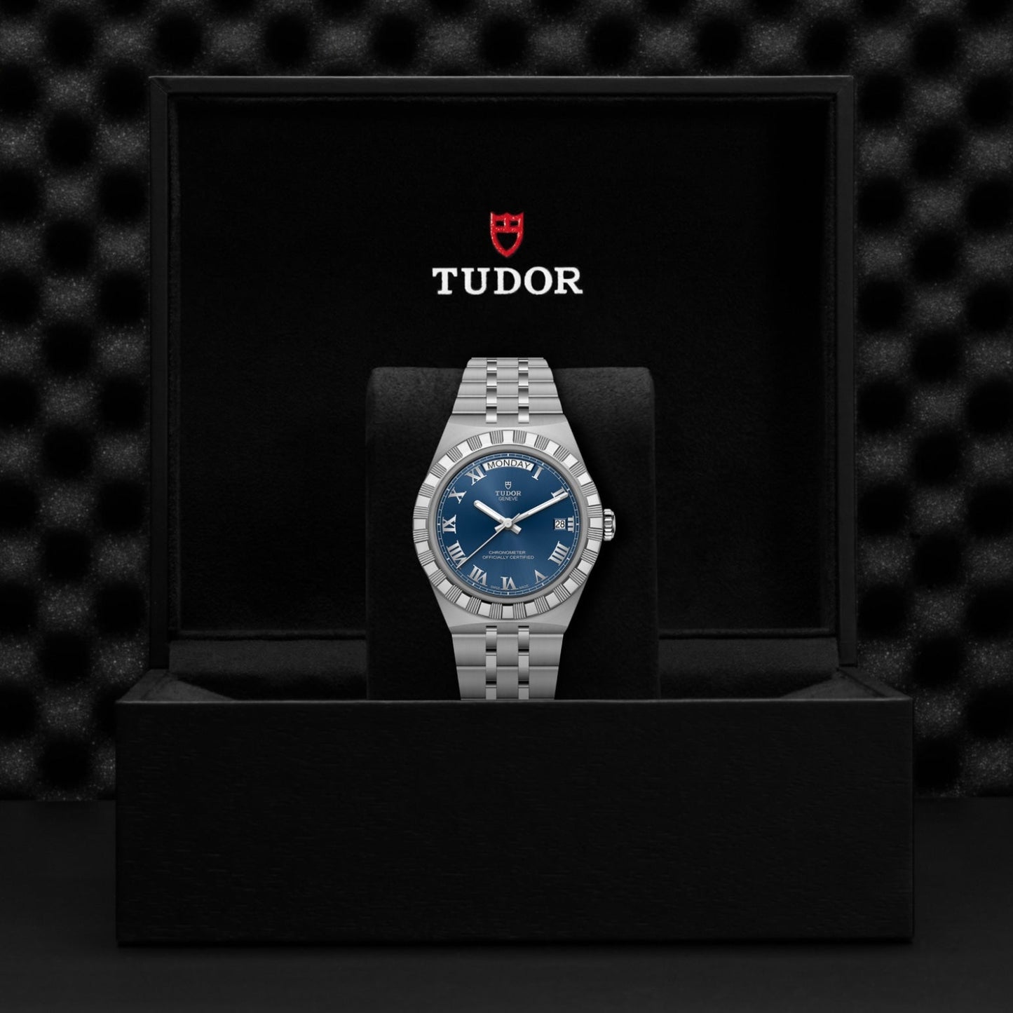 The TUDOR Royal wristwatch, featuring a blue dial and silver metal band, is showcased upright in an open black TUDOR gift box with the logo and red shield emblem inside the lid, highlighting its sport-chic automatic design.