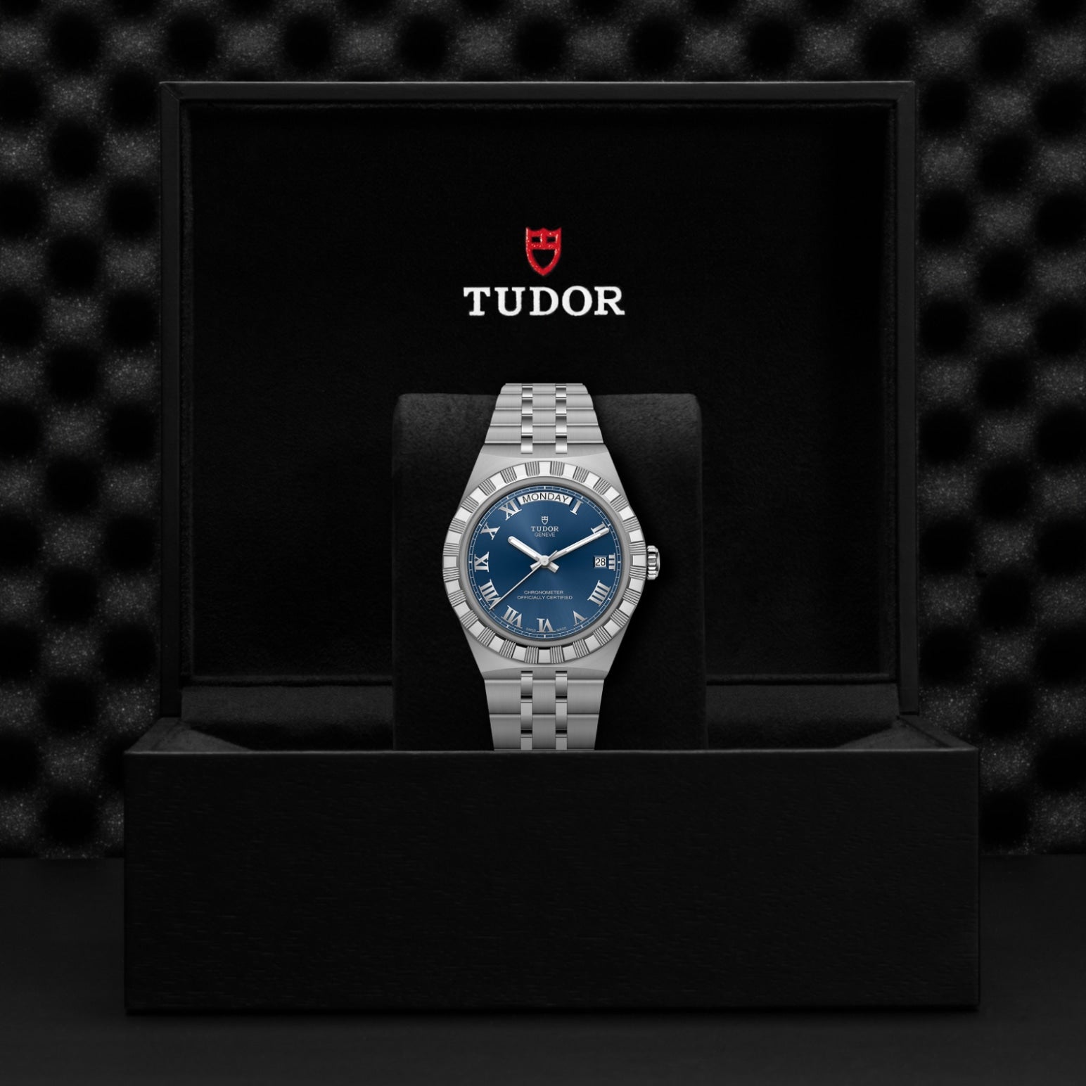 The TUDOR Royal wristwatch, featuring a blue dial and silver metal band, is showcased upright in an open black TUDOR gift box with the logo and red shield emblem inside the lid, highlighting its sport-chic automatic design.