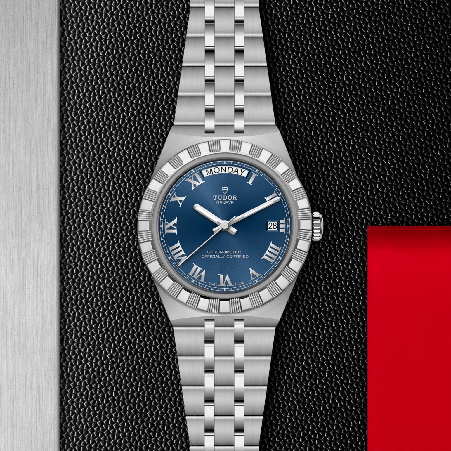 The TUDOR Royal by TUDOR features a silver case, blue dial with Roman numerals, day-date displays, and a metal bracelet—shown on a textured black surface with silver and red accents—a true icon among automatic sport-chic watches.