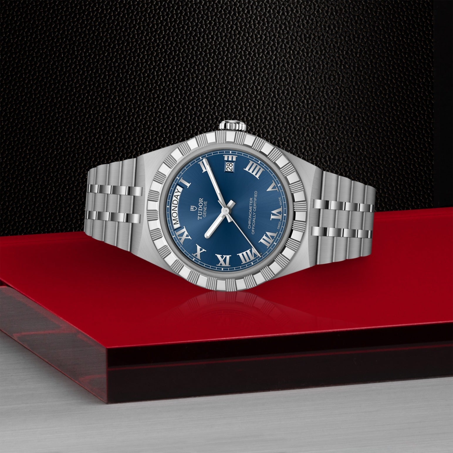 A silver TUDOR Royal wristwatch from TUDOR with a blue Roman numeral dial and day-date display is displayed on a glossy red surface against a black textured background.