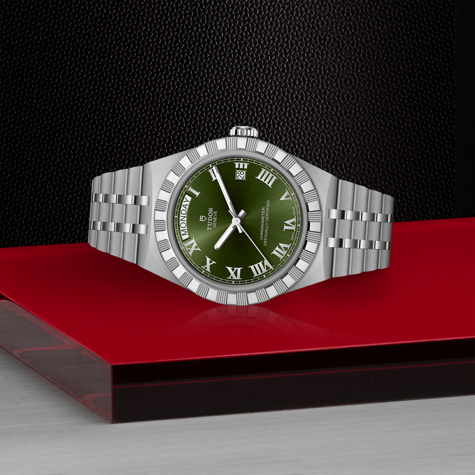 The TUDOR Royal by TUDOR is a luxury silver wristwatch with a green Roman numeral dial, day/date display, and metal bracelet—stylishly presented on a glossy red and black surface for sport-chic watch enthusiasts.