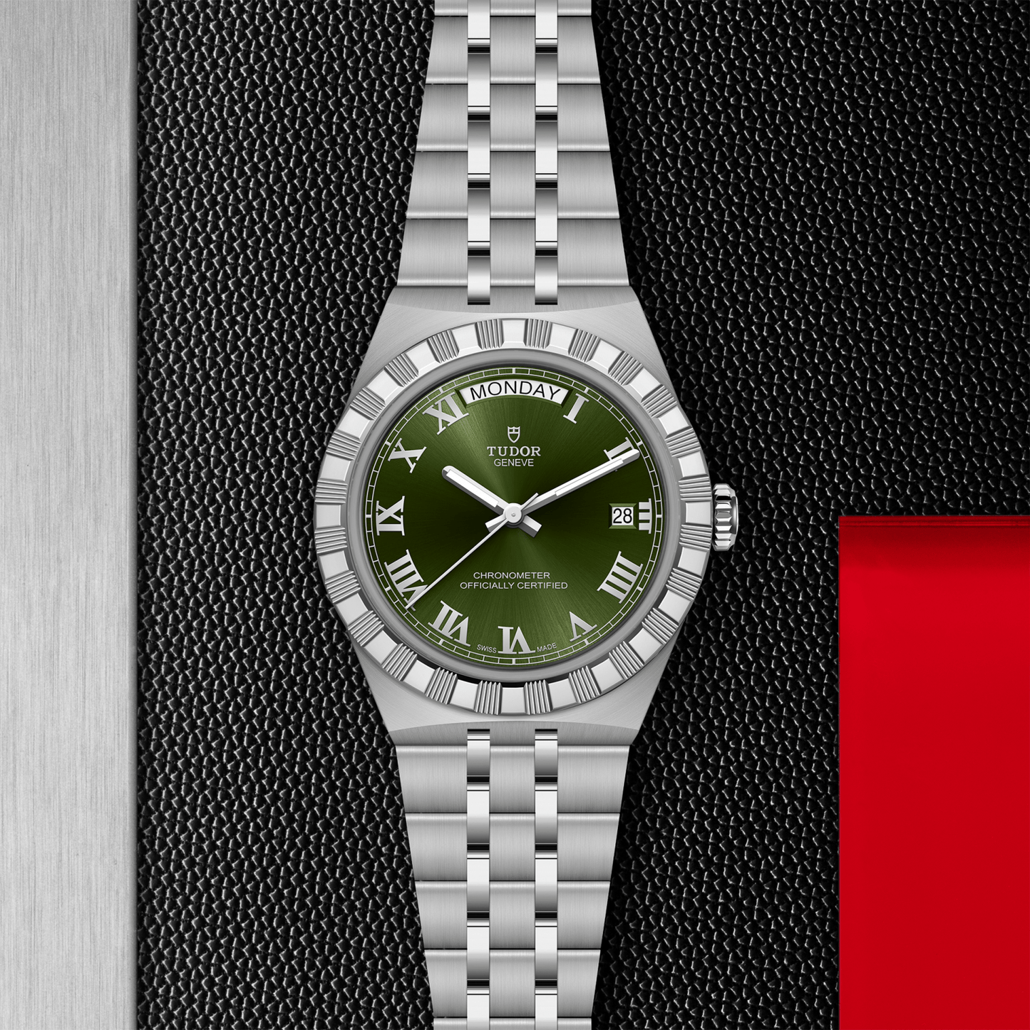 The TUDOR Royal by TUDOR features a stainless steel case, silver metal bracelet, green Roman numeral dial with day and date displays, displayed on a textured black and silver surface accented in red.