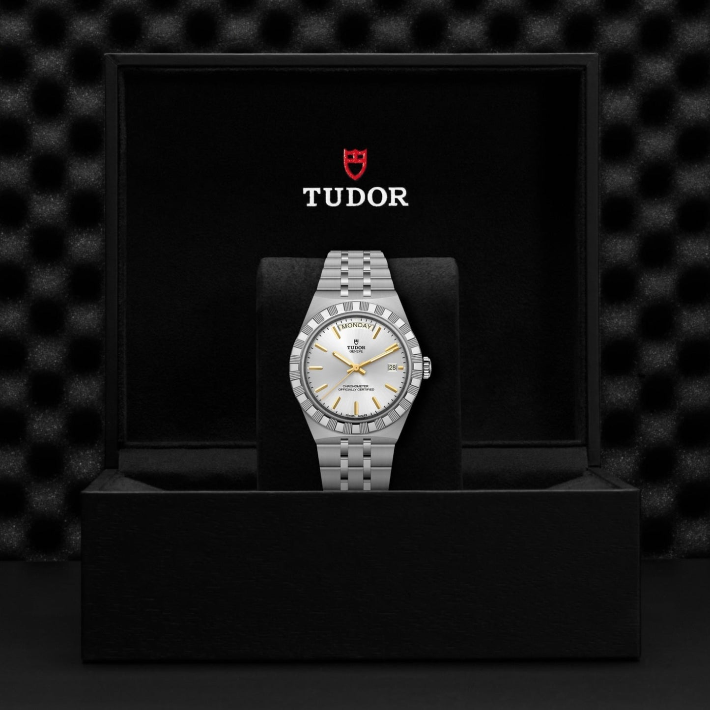 A silver TUDOR Royal wristwatch is displayed upright in an open black box, featuring a white dial with gold-tone markers and the TUDOR logo printed inside the lid, highlighting the refined elegance of this stainless steel watch.