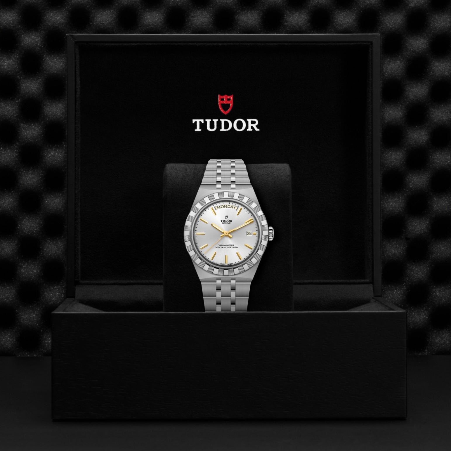 A silver TUDOR Royal wristwatch is displayed upright in an open black box, featuring a white dial with gold-tone markers and the TUDOR logo printed inside the lid, highlighting the refined elegance of this stainless steel watch.
