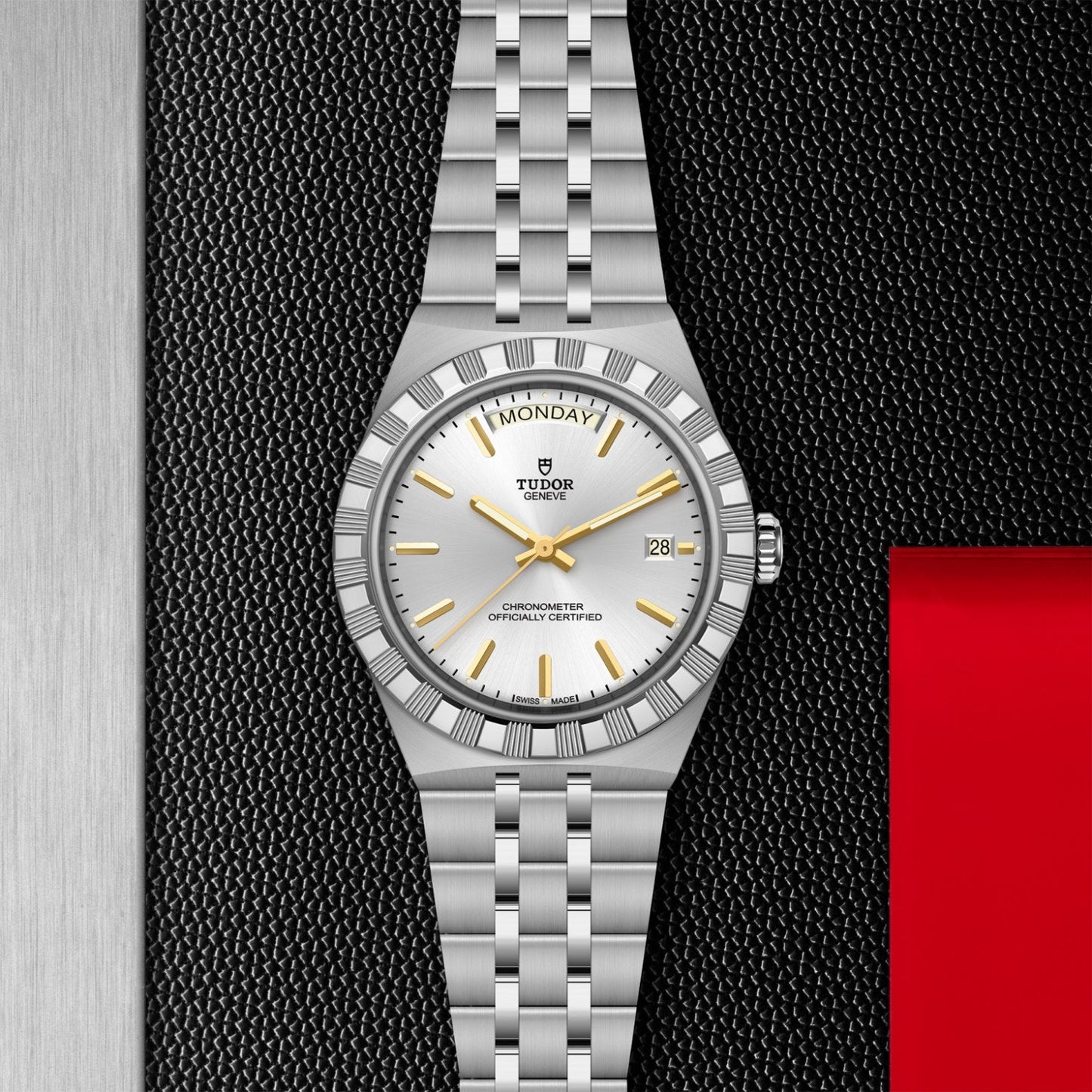 The TUDOR Royal by TUDOR features a silver case, white dial, gold hour markers, and a Jubilee-style bracelet. Displayed against a black, silver, and red background, this automatic watch shows the day at 12 and date at 3 o’clock.