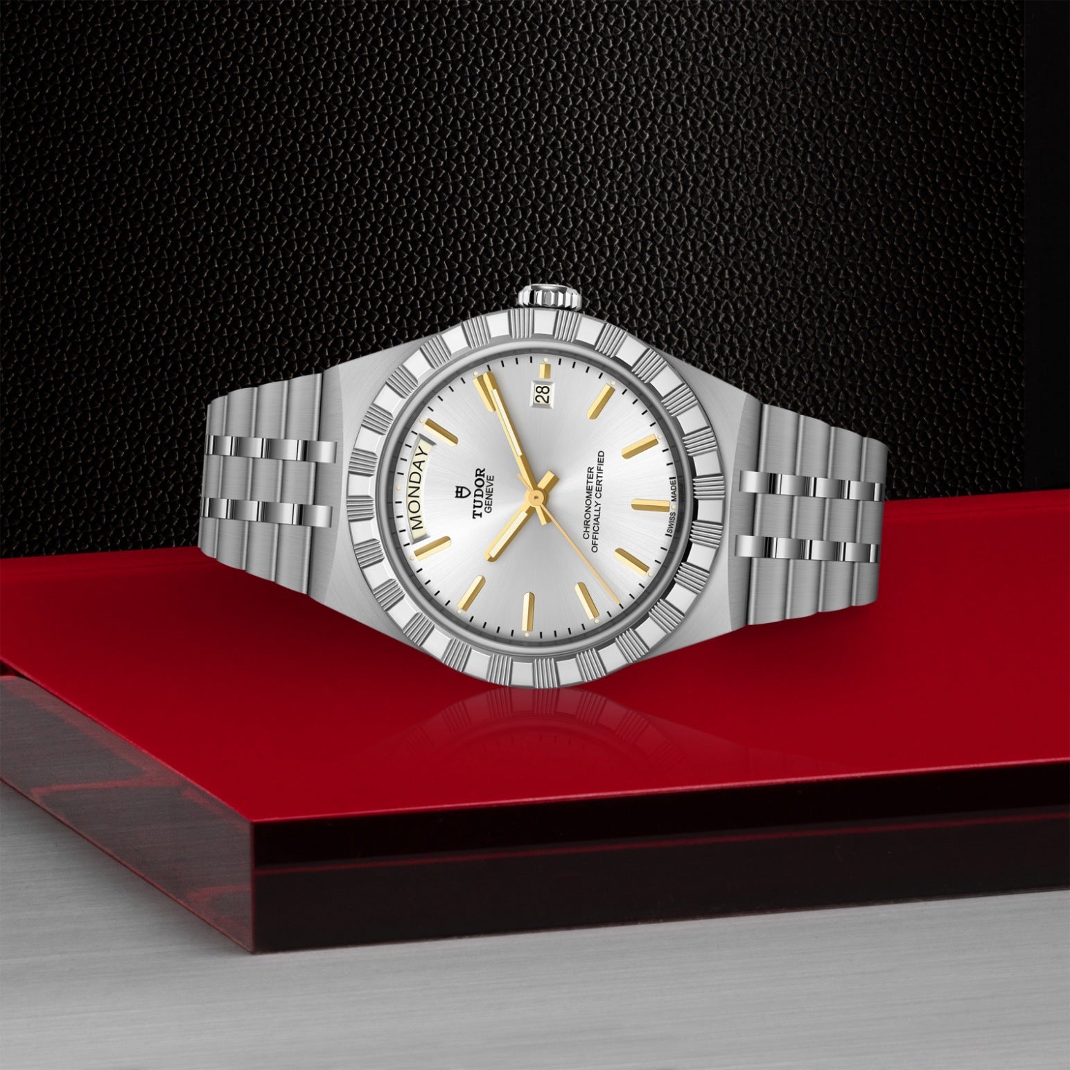 The TUDOR Royal by TUDOR, featuring a silver case, white dial with gold hour markers, and a metal link band, is shown on a red and gray surface against a black textured background. This automatic watch displays the day and date.