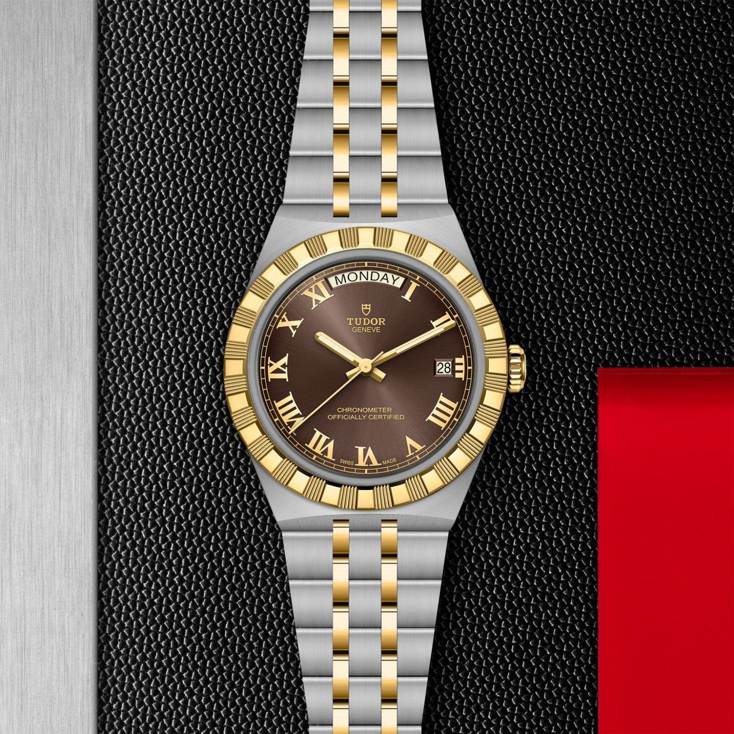 The TUDOR Royal by TUDOR features a two-tone stainless steel and gold bracelet, black dial with gold Roman numerals, day-date display, set against a textured black background with silver and red accents.