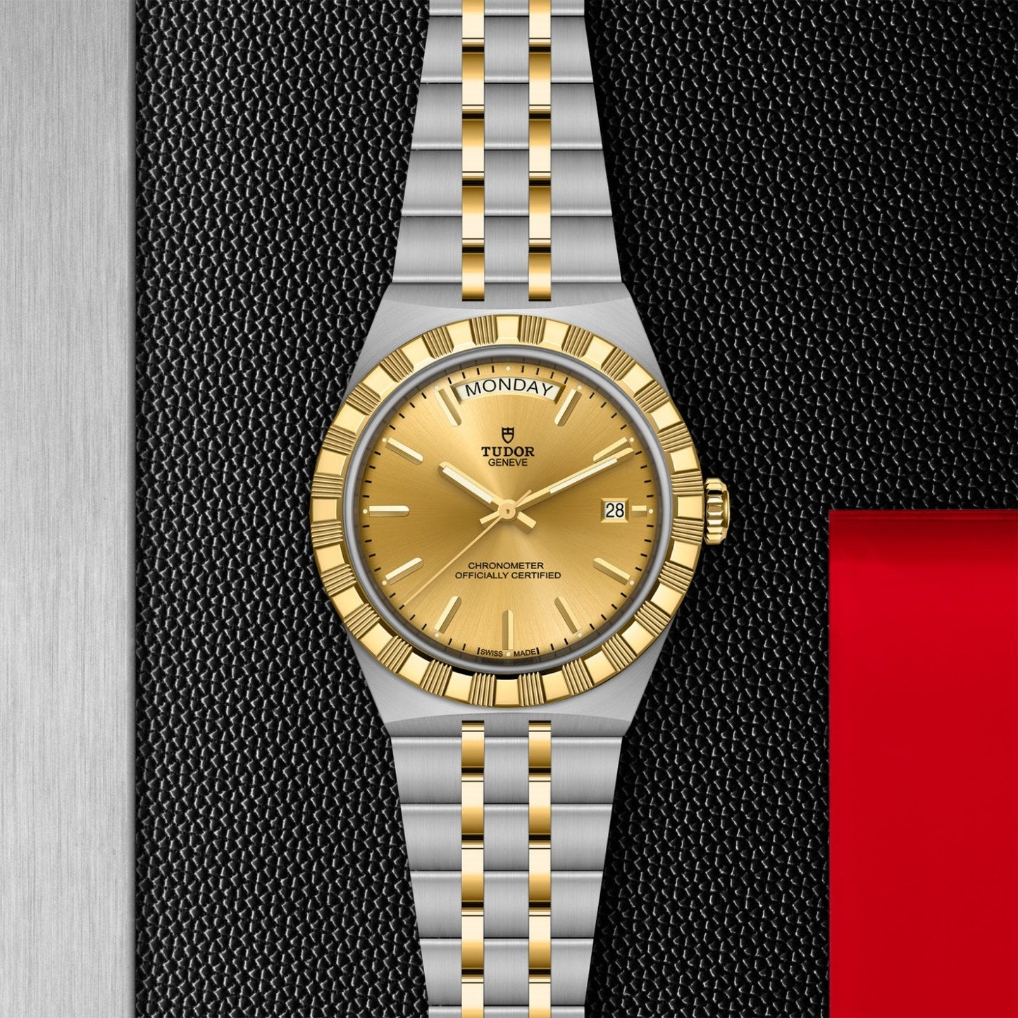 A TUDOR Royal luxury wristwatch by TUDOR, featuring a gold dial, gold fluted bezel, and two-tone silver and gold bracelet, is displayed on a textured black surface beside metallic and red panels.