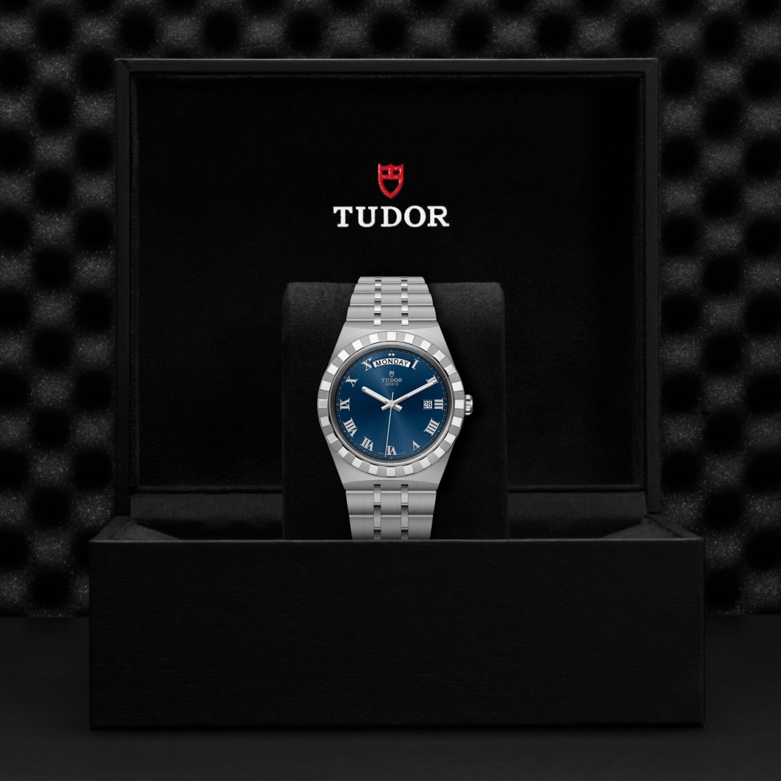 A TUDOR Royal wristwatch with a silver case, blue dial, and Roman numerals is presented upright in an open black box with the TUDOR logo and red shield, set against a textured black background that highlights this automatic watch.