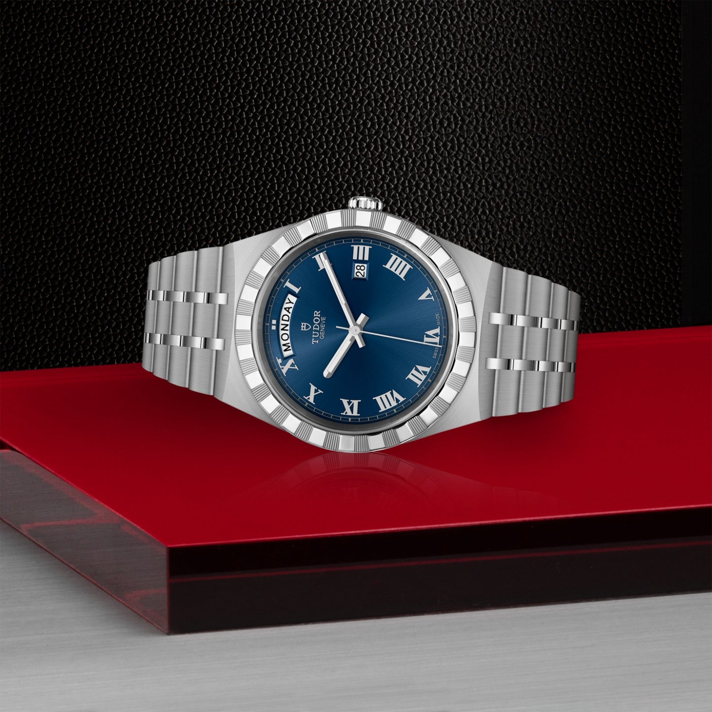 The TUDOR Royal by TUDOR features a silver stainless steel case, blue dial with Roman numerals, and day-date display. Displayed on a red platform against a textured black background, it's perfect for automatic sport-chic watch fans.