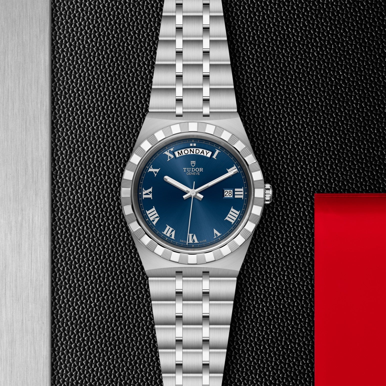 The TUDOR Royal by TUDOR features a blue dial with Roman numerals and day-date display, set on a textured black surface beside brushed metal and a red accent, reflecting its automatic sport-chic design.