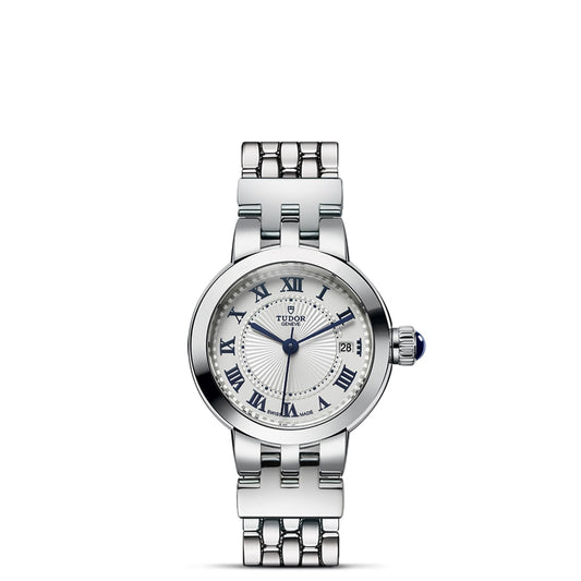 The TUDOR Clair de Rose is a silver wristwatch with a round opaline dial, blue lacquered Roman numerals, date window at 3 o'clock, blue hands, metal link bracelet, and a crown with a striking blue accent.