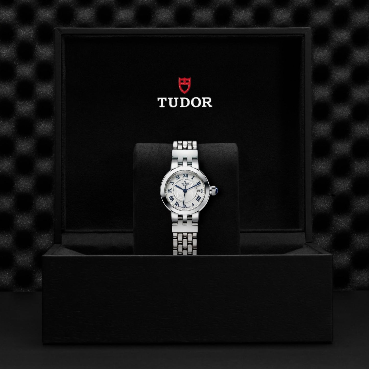 The TUDOR Clair de Rose wristwatch, featuring a metallic bracelet and opaline dial, is showcased in a black presentation box with the TUDOR logo and red shield, set against a textured black background.