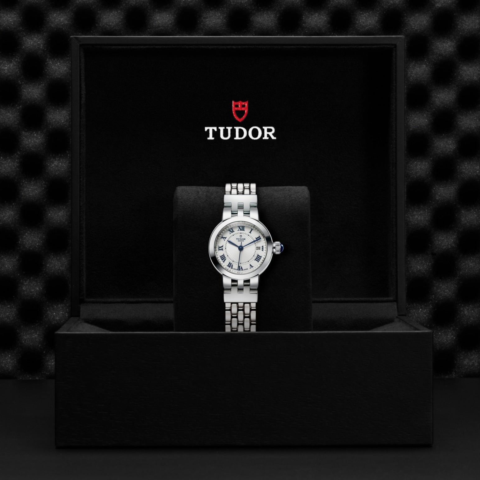 The TUDOR Clair de Rose wristwatch, featuring a metallic bracelet and opaline dial, is showcased in a black presentation box with the TUDOR logo and red shield, set against a textured black background.