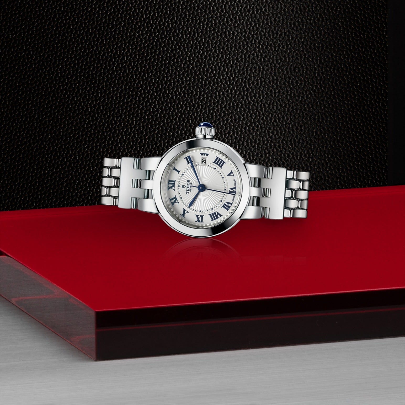 A TUDOR Clair de Rose watch with blue lacquered Roman numerals on an opaline dial and a stainless steel bracelet is elegantly displayed on a glossy red and gray surface against a textured black background.
