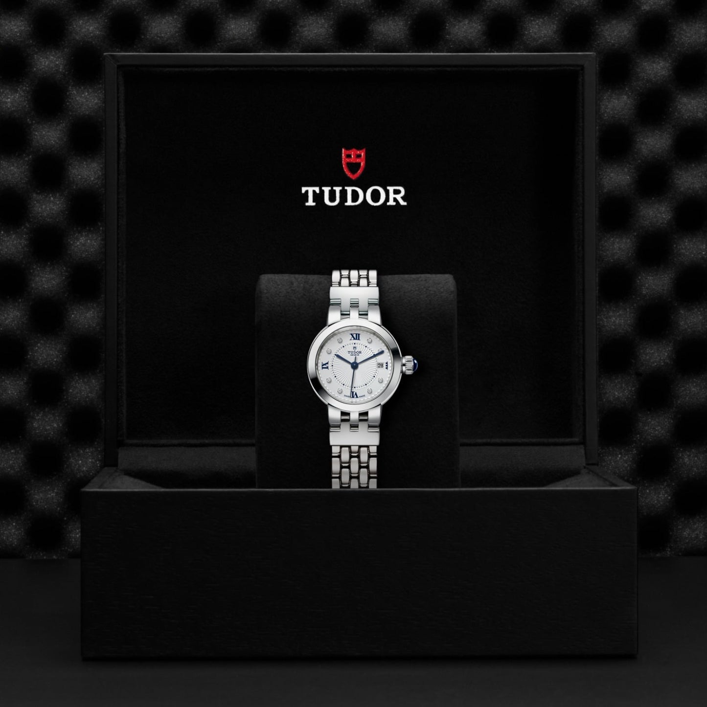 A TUDOR Clair de Rose wristwatch with an opaline dial and blue Roman numerals is displayed upright on a black cushioned stand inside an open black TUDOR box, the brand logo visible on the inner lid.