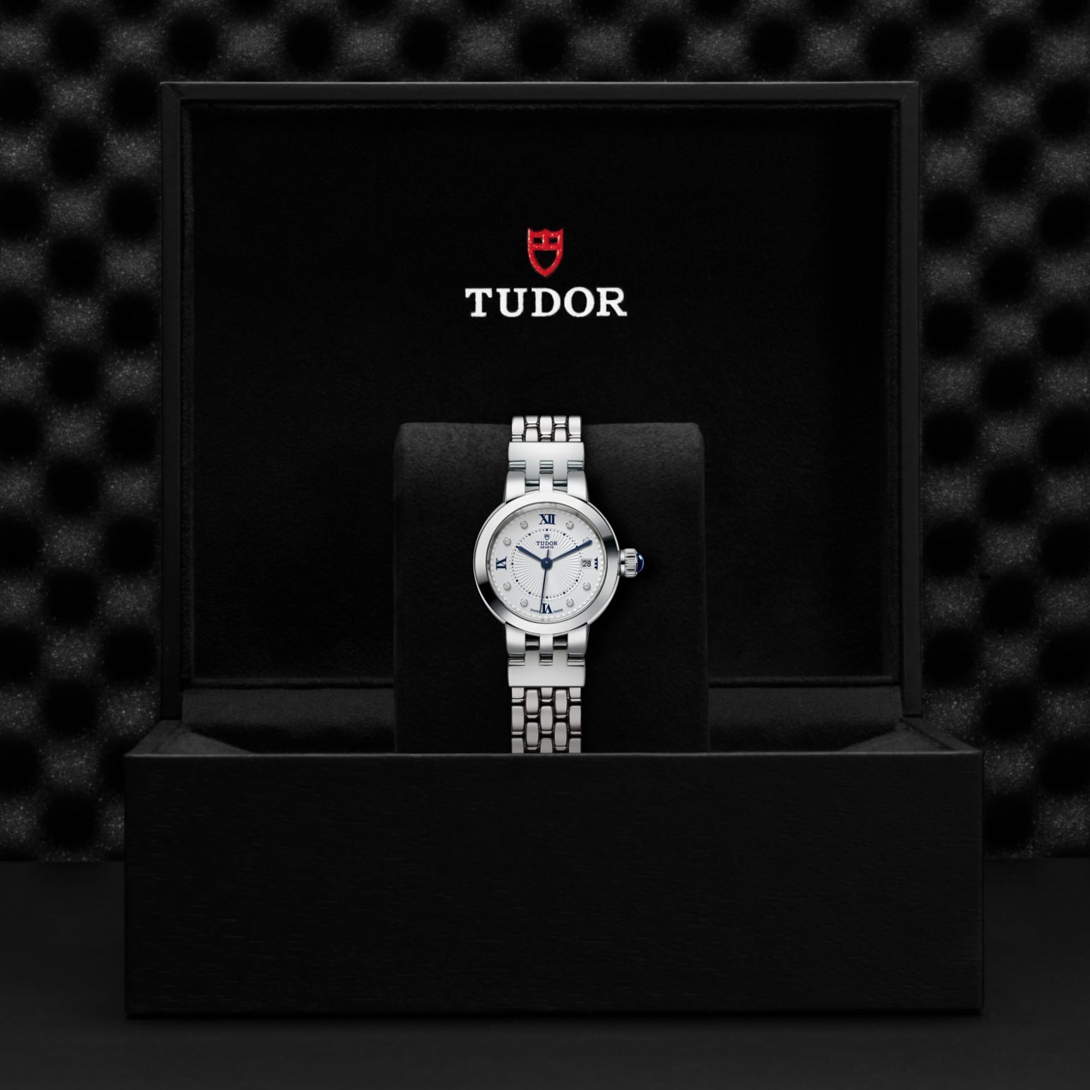 A TUDOR Clair de Rose wristwatch with an opaline dial and blue Roman numerals is displayed upright on a black cushioned stand inside an open black TUDOR box, the brand logo visible on the inner lid.
