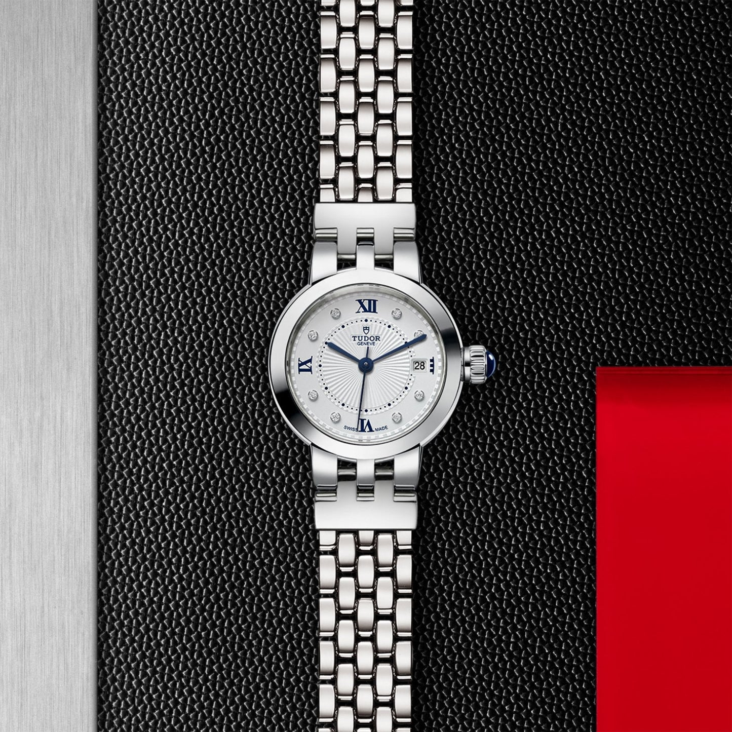 The TUDOR Clair de Rose wristwatch features an opaline dial, blue hands, blue lacquered Roman numerals, and a date window, shown on a textured black background with silver and red accents.