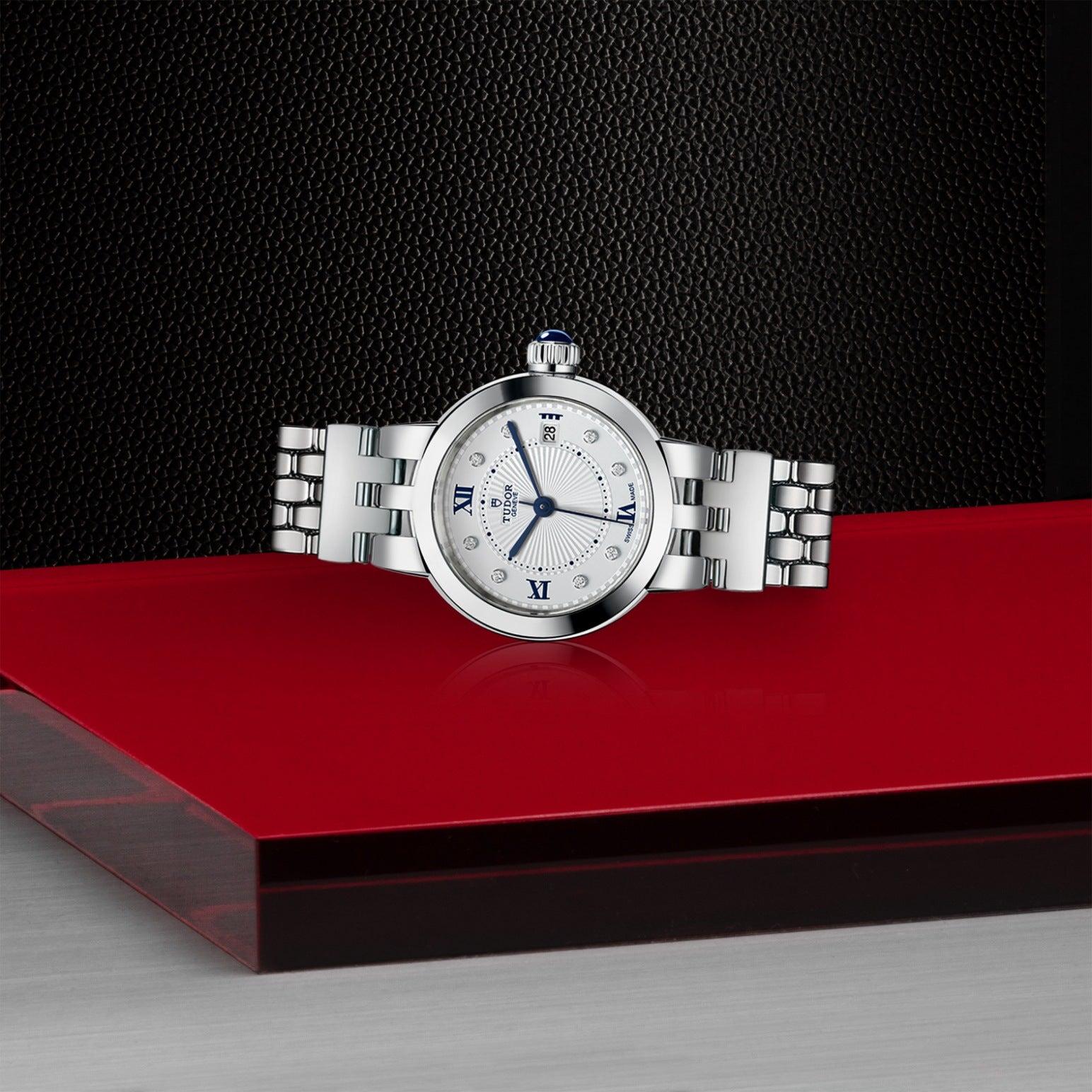 The TUDOR Clair de Rose, a silver luxury wristwatch with a white face, blue lacquered Roman numerals, and a metal bracelet, rests on a glossy red and gray platform against a textured black background.