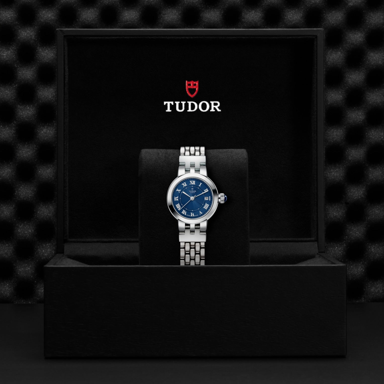 A silver TUDOR Clair de Rose watch with an opaline dial and blue lacquered Roman numerals rests on a black cushion inside an open black TUDOR box displaying the brand’s name and logo on the interior lid.