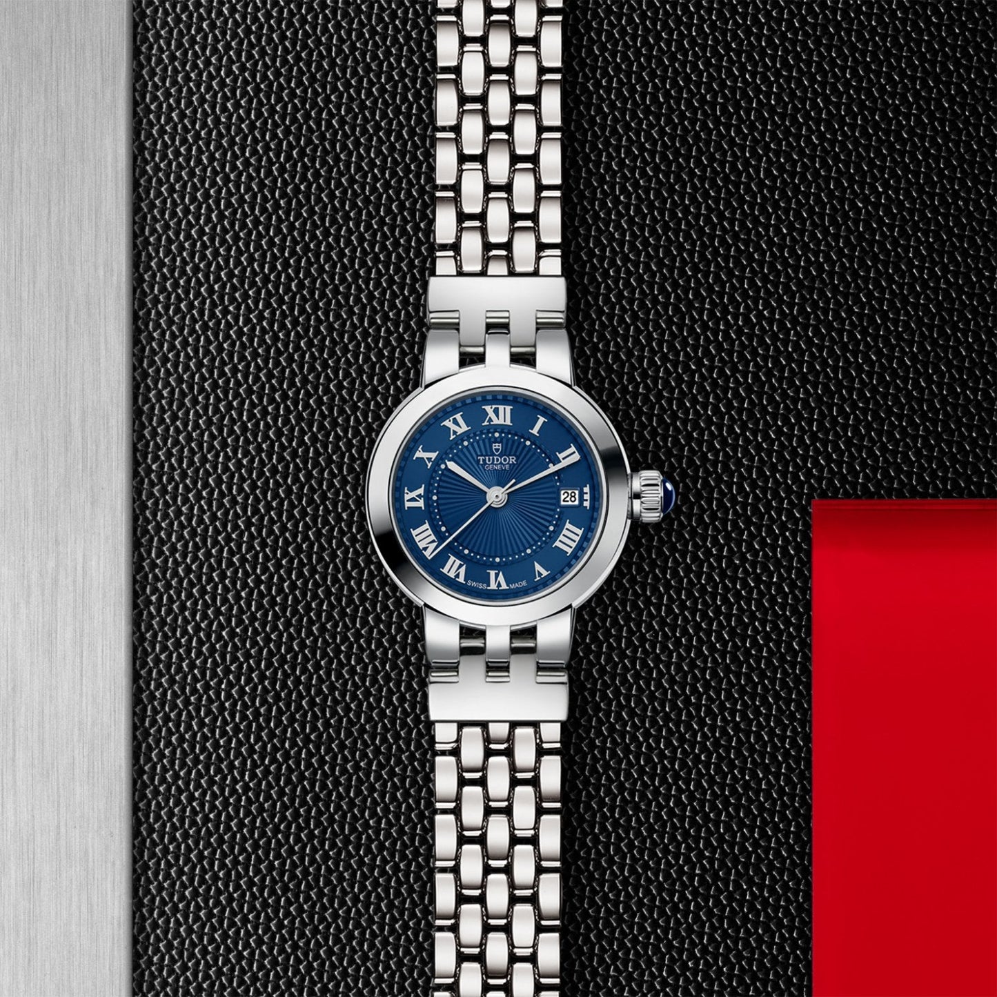 The TUDOR Clair de Rose wristwatch features a blue dial with lacquered Roman numerals and a metal link band, displayed on a textured black surface beside silver and red accents.