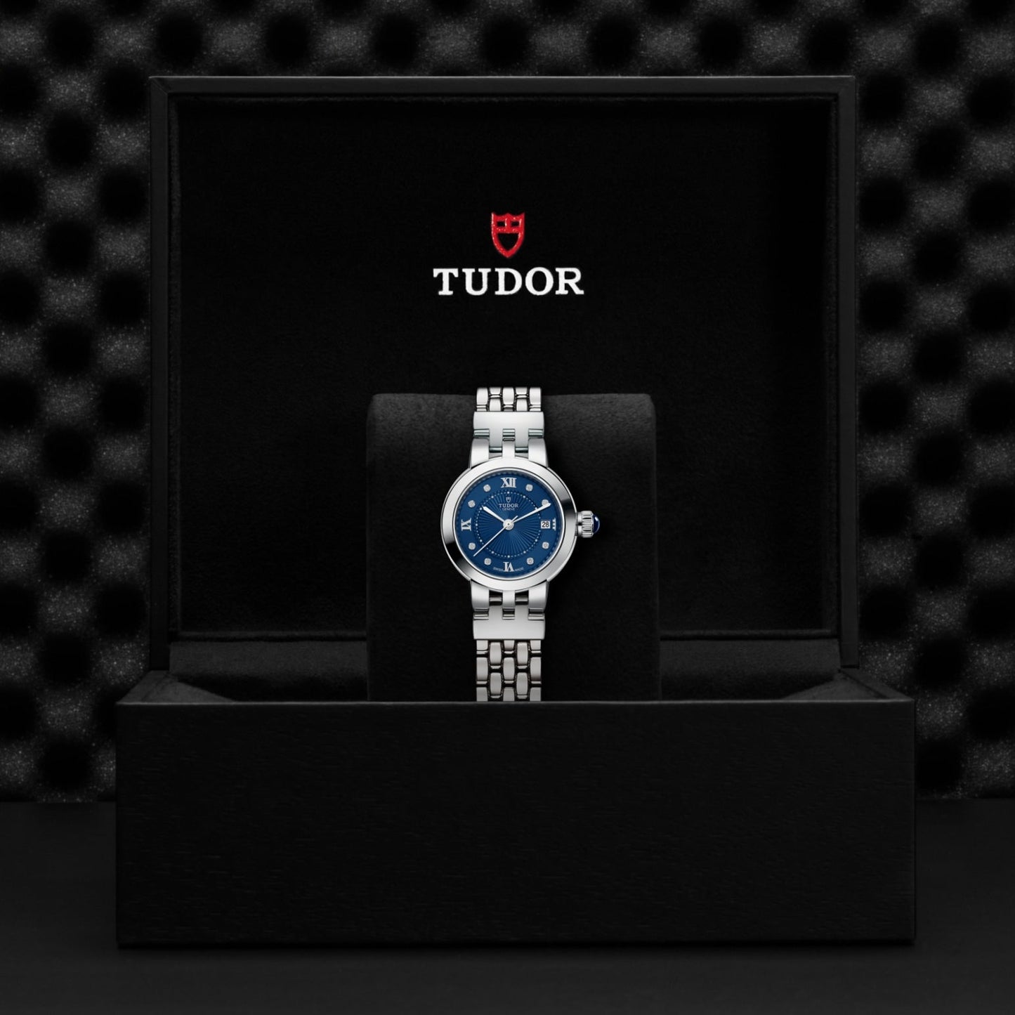A TUDOR Clair de Rose wristwatch with a blue dial and blue Roman numerals is displayed upright in a black presentation box featuring the TUDOR logo and shield emblem in white and red on the inside lid, set against a textured black background.