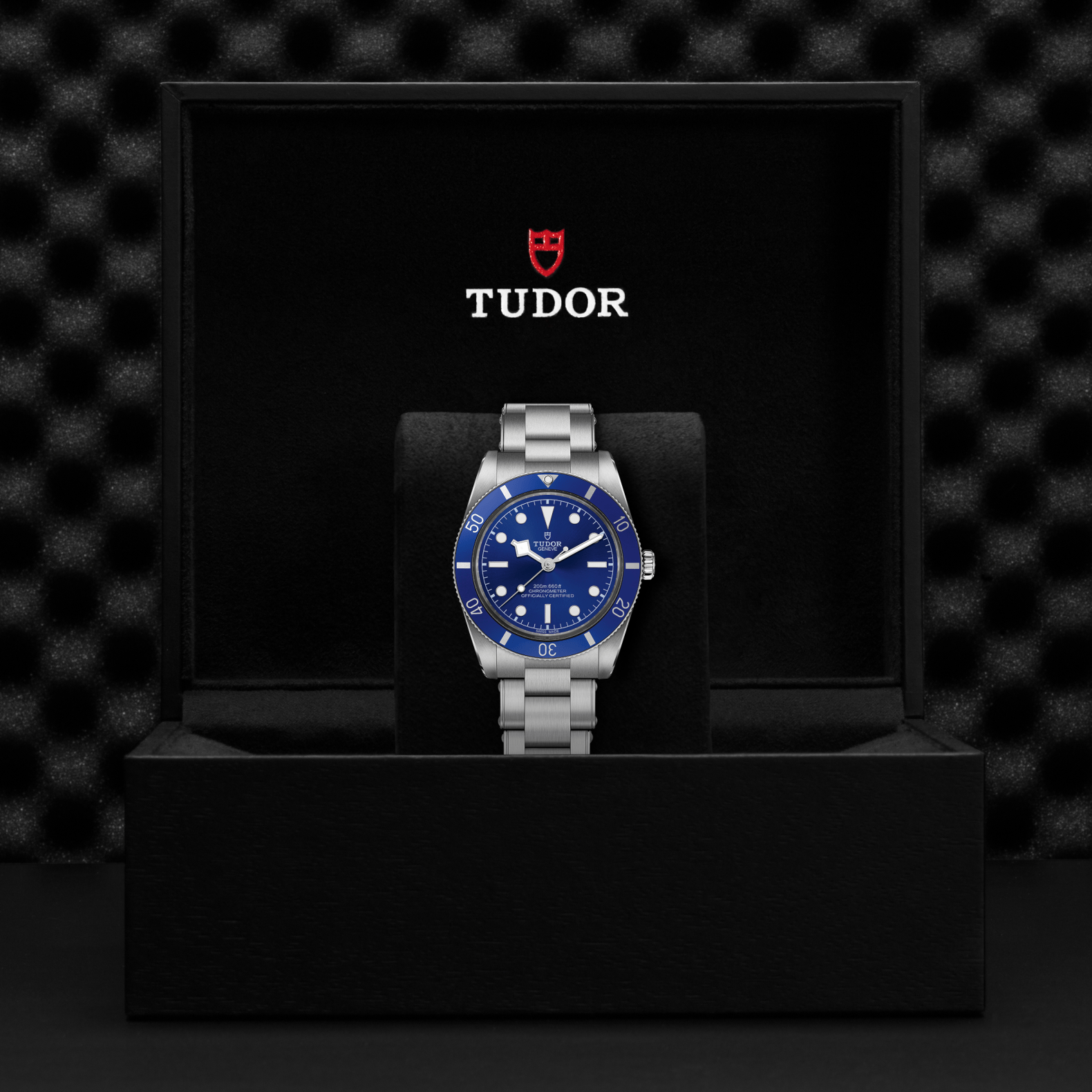 A TUDOR Black Bay 54 dive watch with a Lagoon Blue dial and silver metal band stands upright in a black Tudor box featuring the red shield logo, set against a textured black background that highlights its refined style.