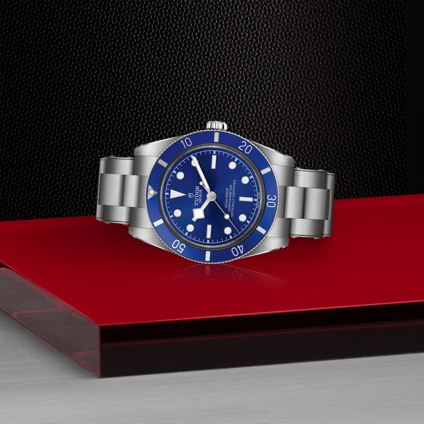 A TUDOR Black Bay 54 dive watch with a Lagoon Blue bezel and dial sits on a glossy red surface, silver case gleaming against a textured black background. The TUDOR timepiece displays 10:10.