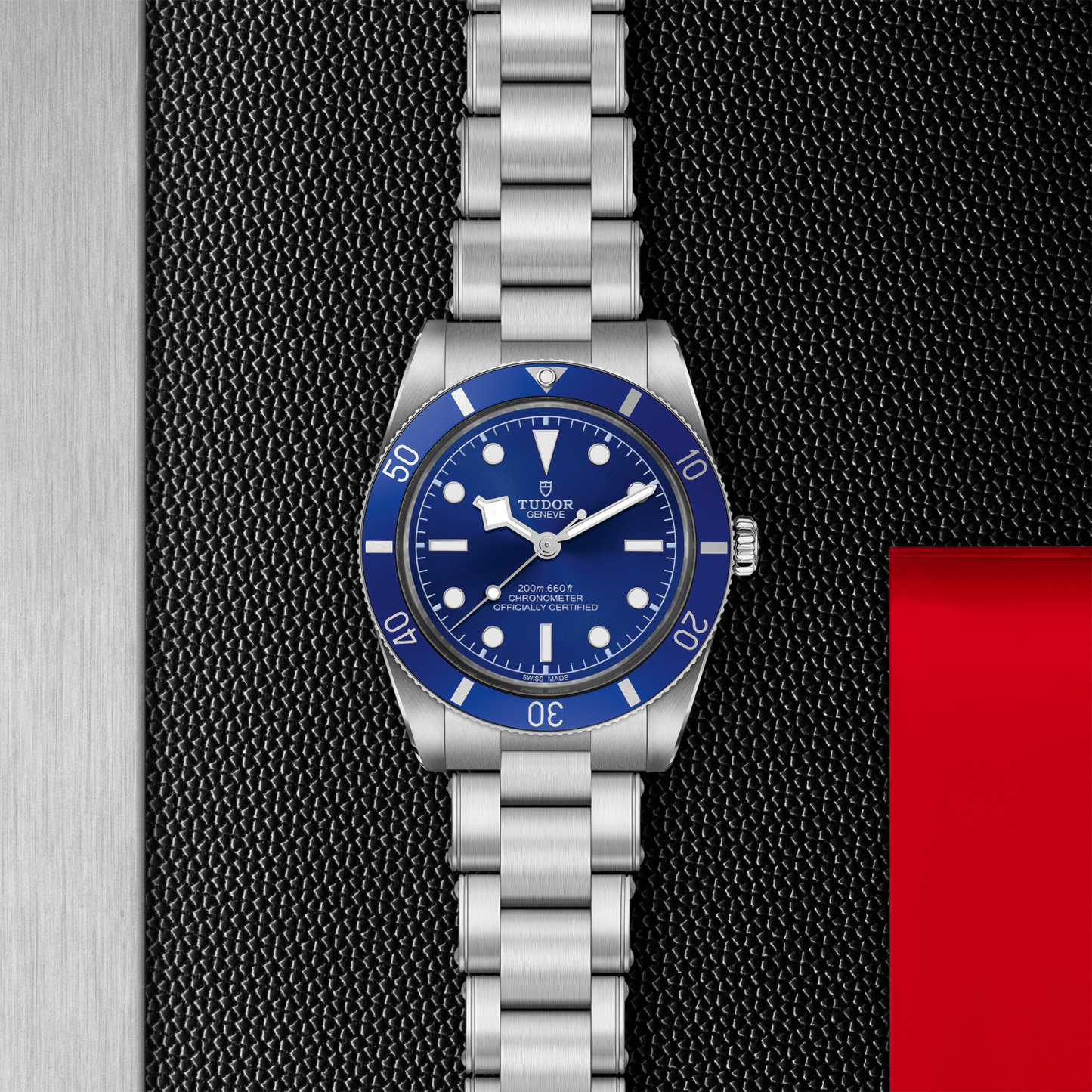 The TUDOR Black Bay 54 dive watch, featuring a Lagoon Blue bezel and dial, is displayed on a textured black background with a red accent on the right and a metallic area on the left.