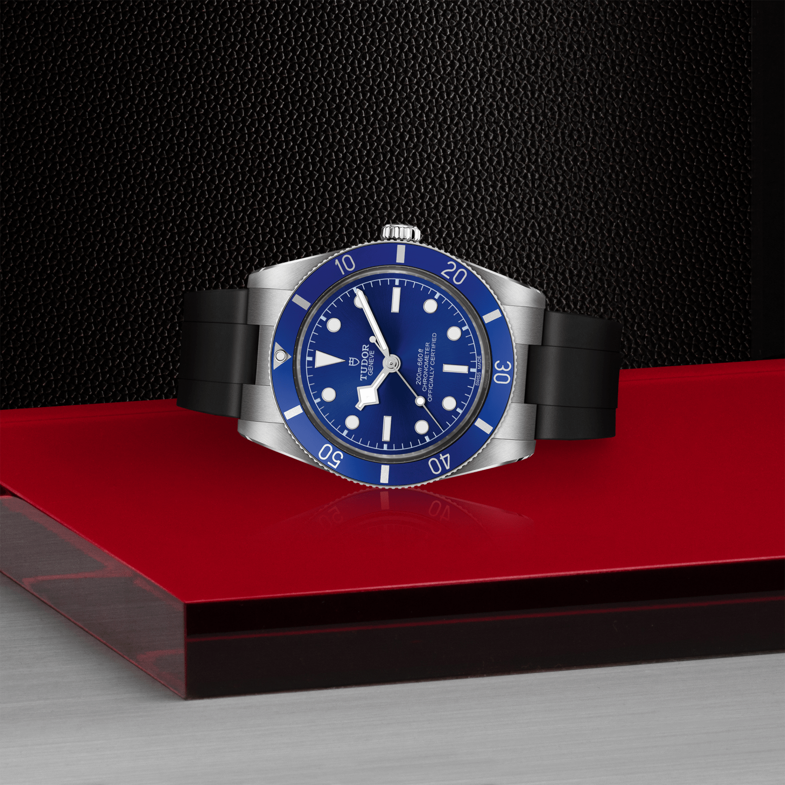 The TUDOR Black Bay 54 dive watch, featuring a Lagoon Blue face and bezel on a black rubber strap, is displayed on a glossy red surface with textured black and metallic backgrounds.