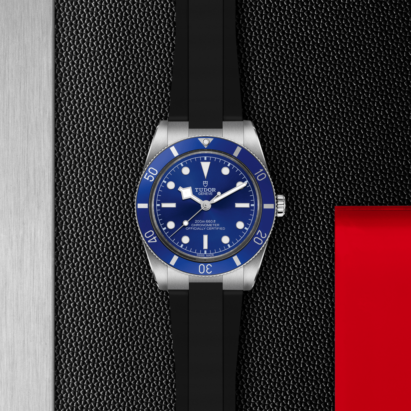 A TUDOR Black Bay 54 dive watch, featuring a Lagoon Blue bezel and dial, silver case, and black rubber strap by TUDOR, is showcased on a textured black and gray background with a vivid red accent on the right.