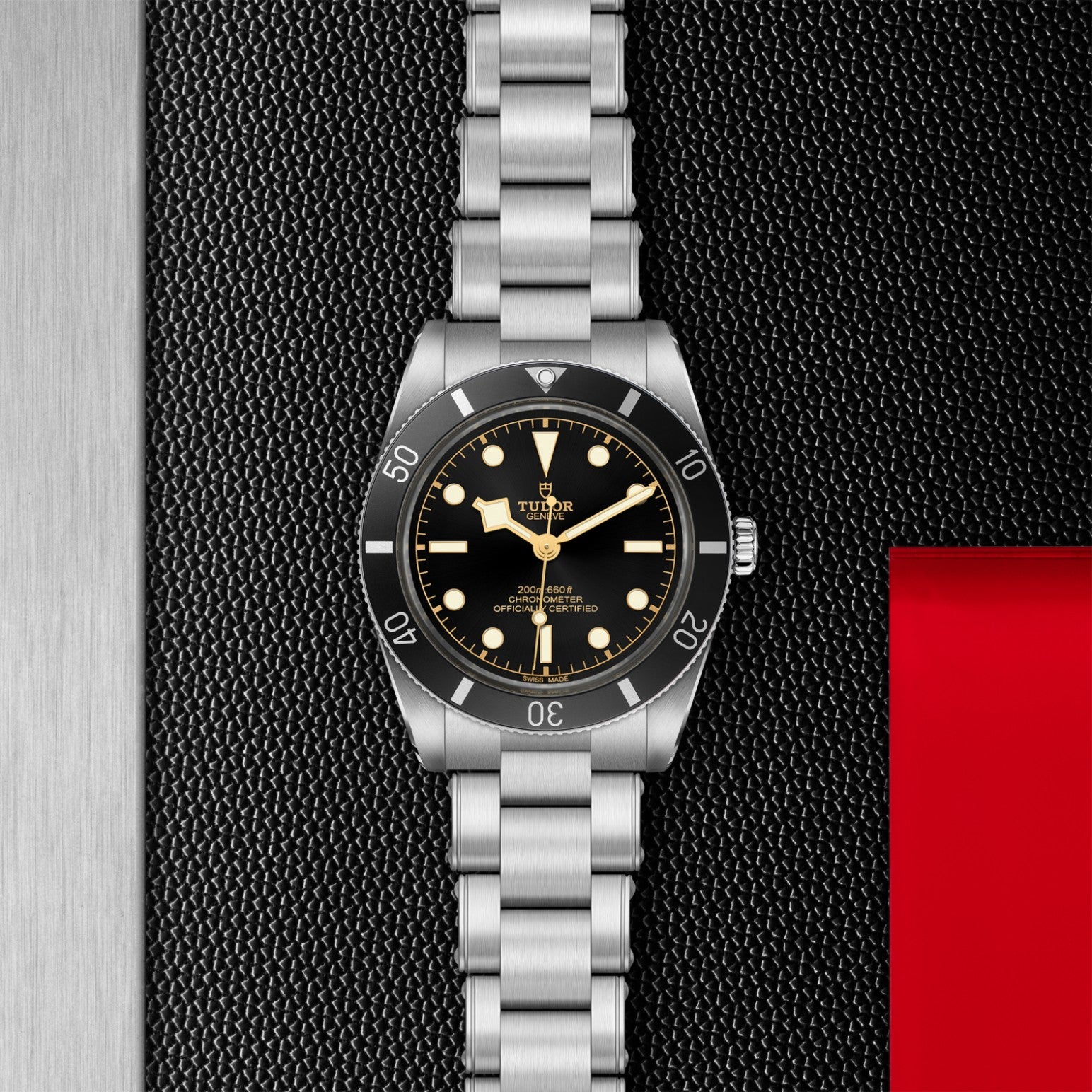 A TUDOR Black Bay 54 dive watch with a black dial and 37mm steel case lies on a textured black surface, set against red and silver accents.