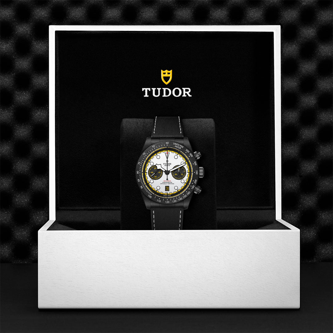 The TUDOR Black Bay Chrono wristwatch, featuring a black strap and a white face with yellow accents, is presented in an open white and black TUDOR box against a textured backdrop.