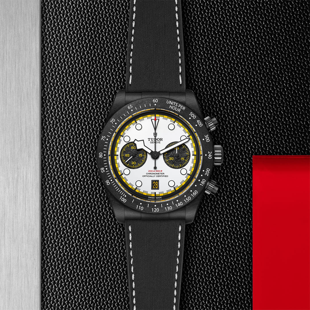 A TUDOR Black Bay Chrono watch featuring a white dial, yellow details, and black leather strap is displayed on a dark textured surface with silver and red accents.