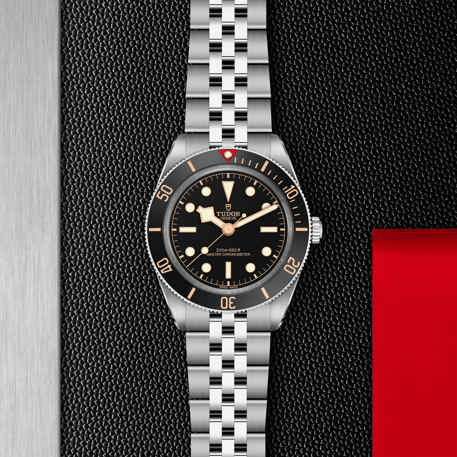 The TUDOR Black Bay 58 dive watch features a stainless steel bracelet, black bezel and dial with luminous markers, shown on a textured black surface with metallic and red accents.