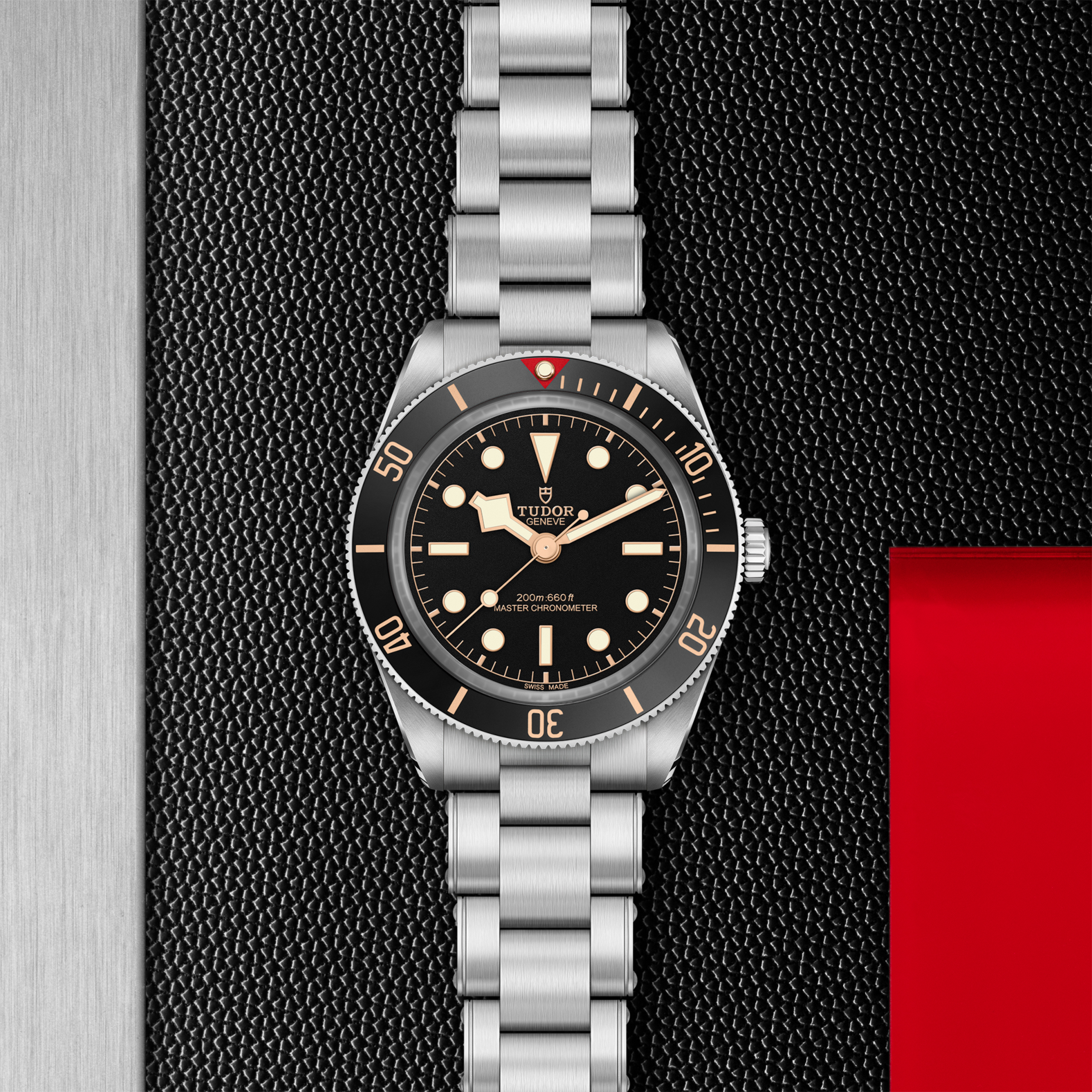 The TUDOR Black Bay 58, featuring a black bezel and dial, luminous markers, and a stainless steel bracelet, is displayed on a textured black surface beside a bold red and metallic background.