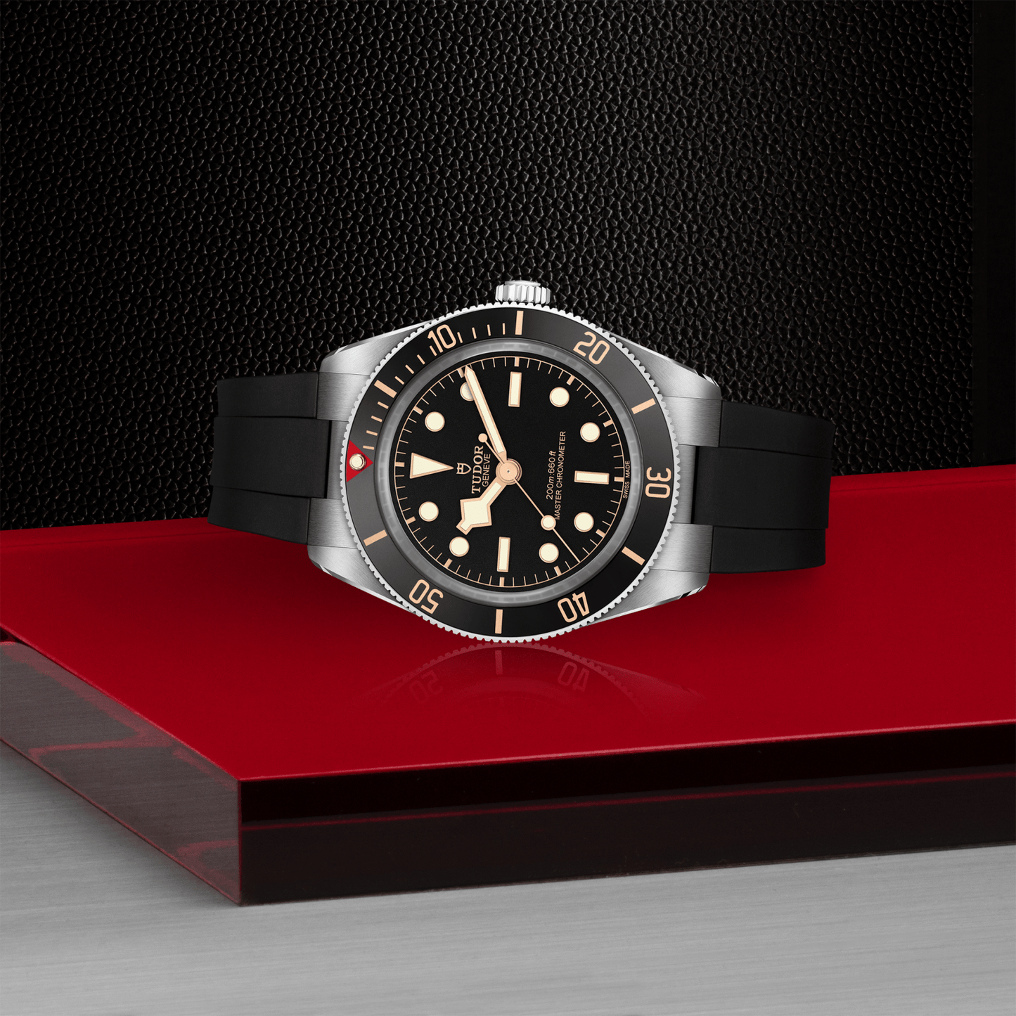 The TUDOR Black Bay 58, a luxury dive watch with a black dial, silver bezel, and black rubber strap, is elegantly displayed on a glossy red and silver surface against a textured black background—a true statement piece from TUDOR.