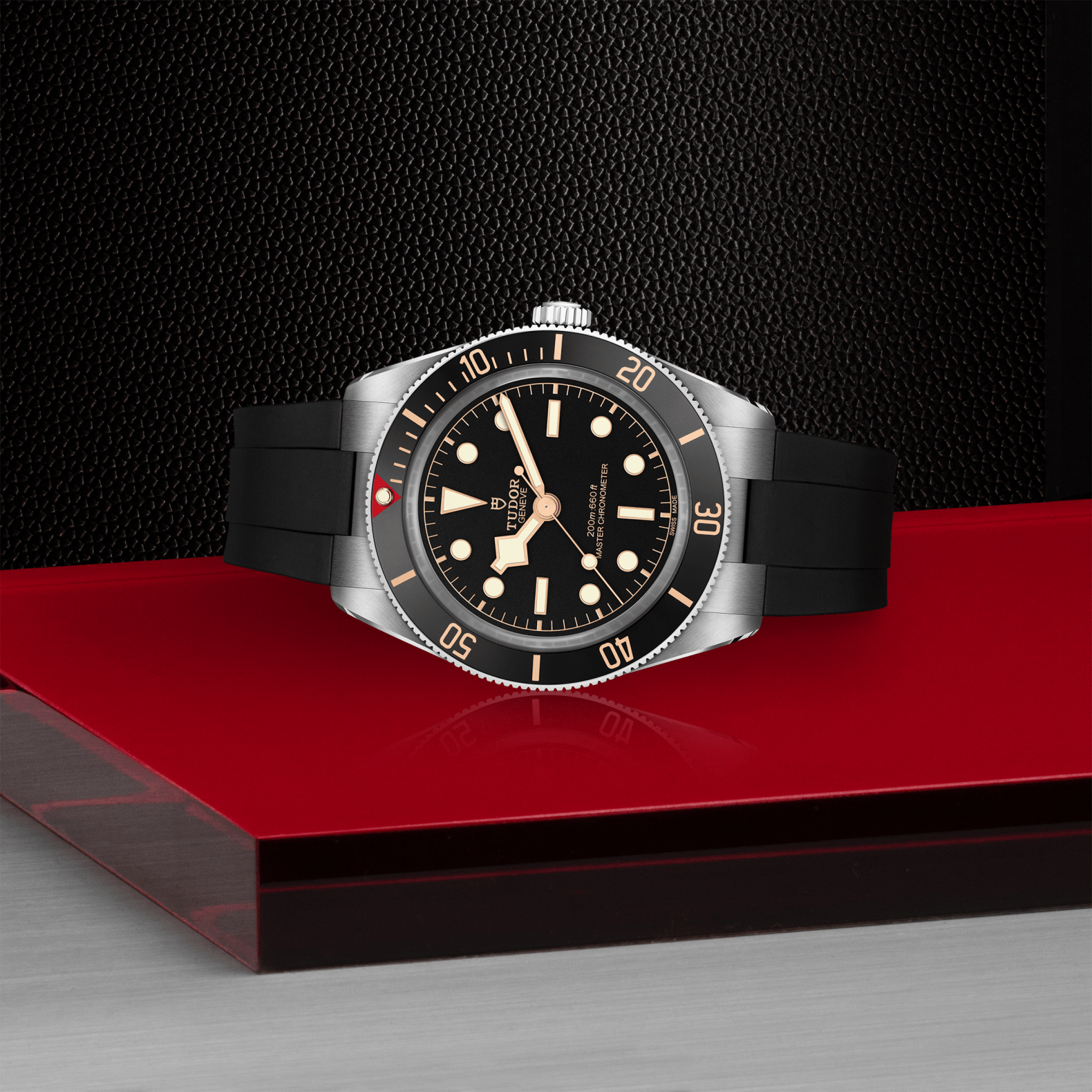 The TUDOR Black Bay 58, a luxury dive watch with a black dial, silver bezel, and black rubber strap, is elegantly displayed on a glossy red and silver surface against a textured black background—a true statement piece from TUDOR.